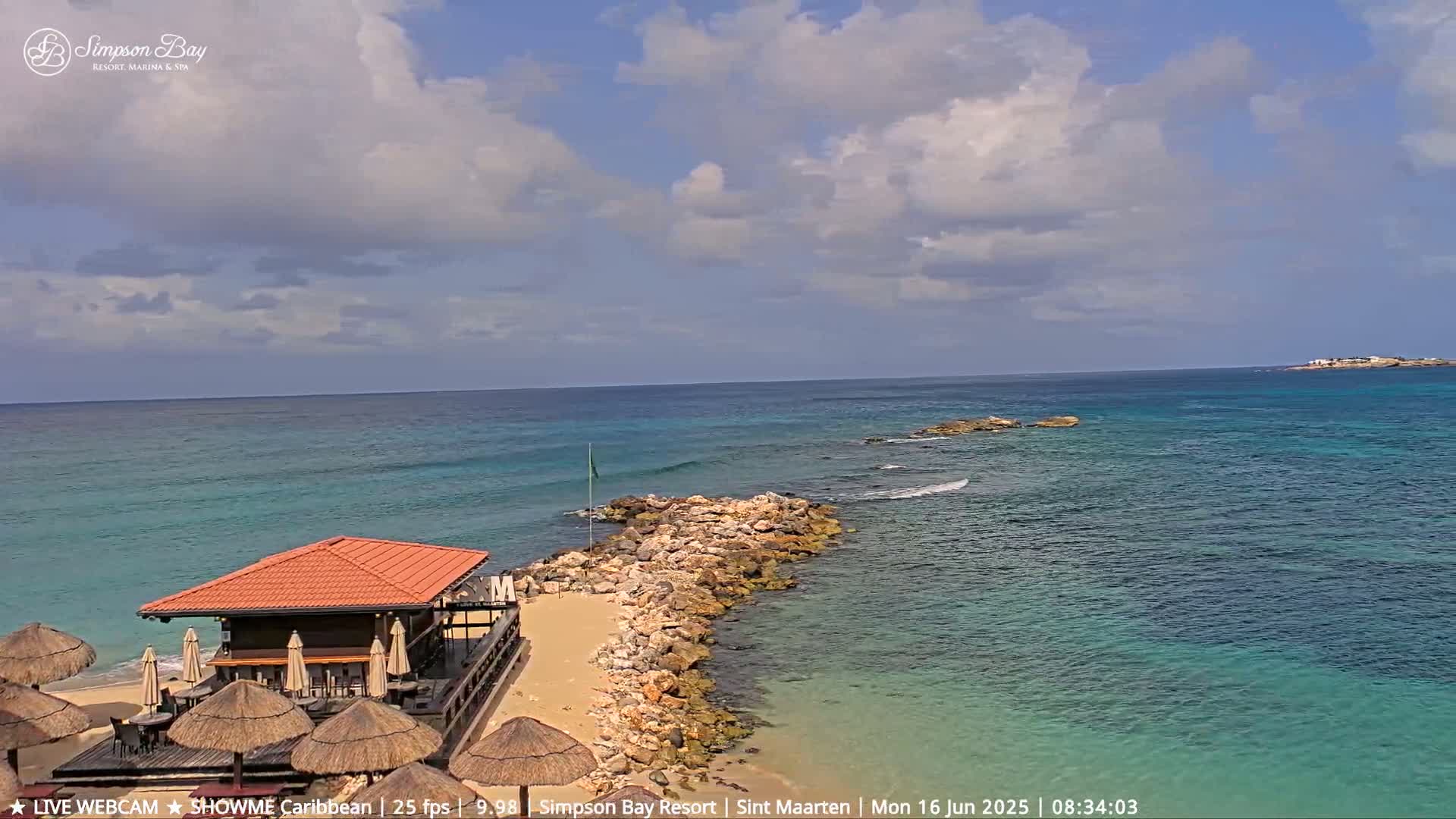 A partly cloudy day shows a beach bar on a sandy shore with a rocky jetty extending into teal-colored water, toward a small island in the distance.