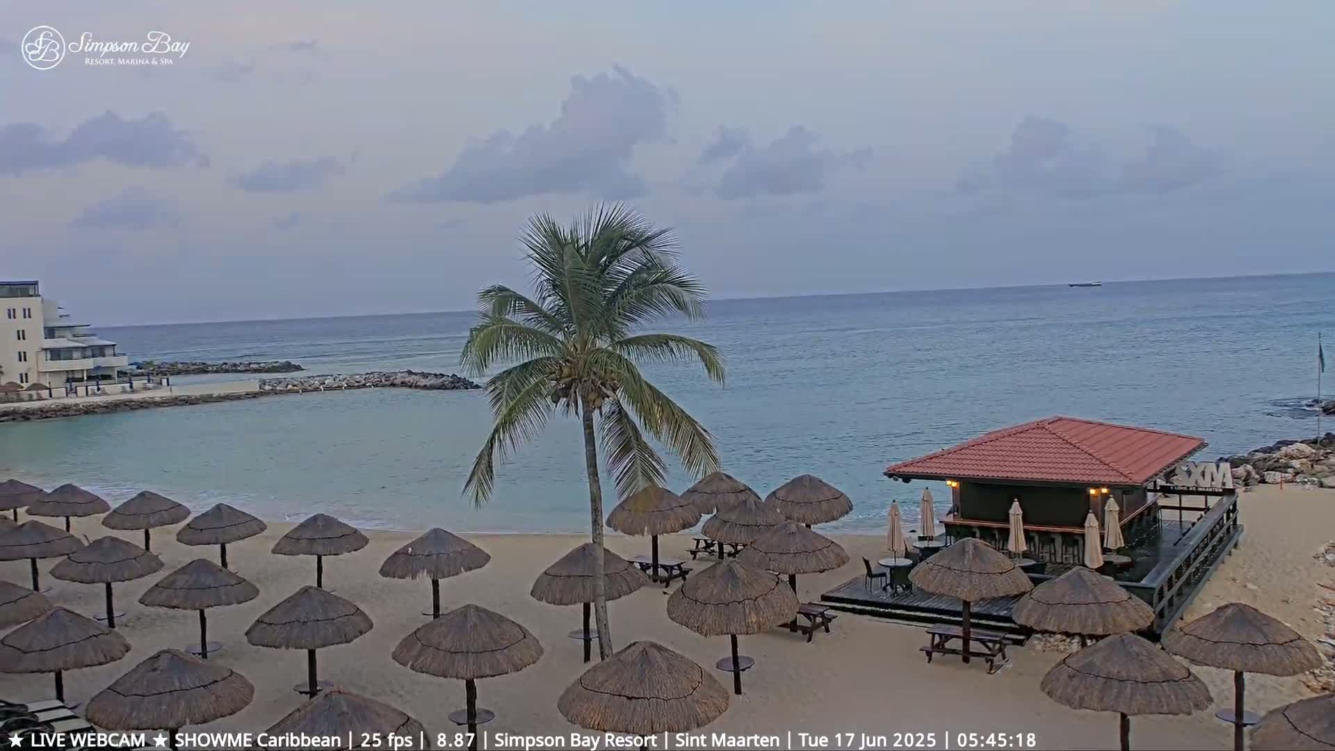 A sandy beach with numerous thatched umbrellas and a beach bar is shown under a partly cloudy sky, overlooking a calm ocean.