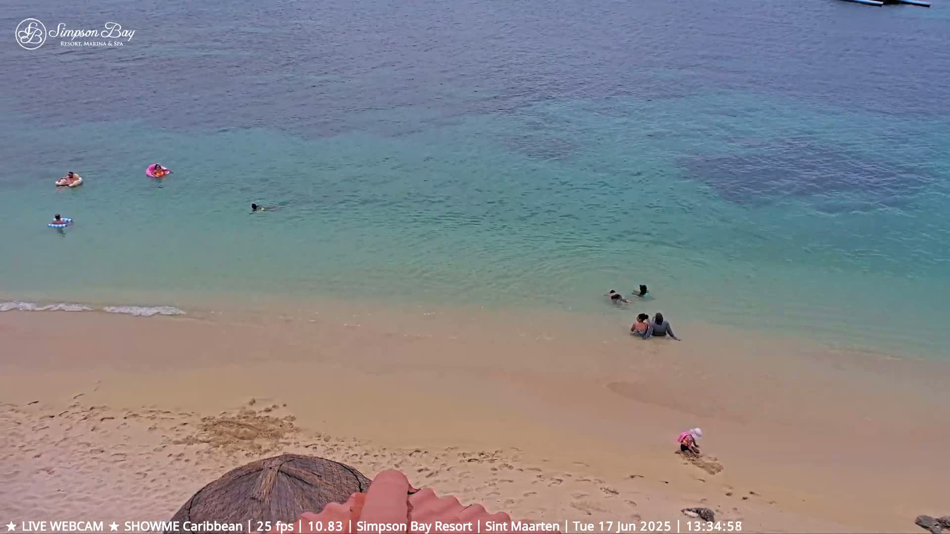 A sandy beach with calm, clear turquoise water shows several people swimming and playing near the shore.