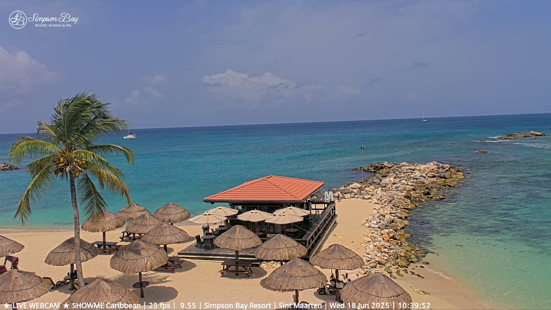 A beach bar with thatched umbrellas sits on a sandy beach next to a rocky outcropping, overlooking a calm turquoise ocean under a mostly sunny sky.