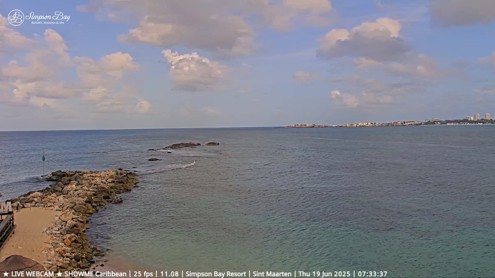 A rocky shoreline extends into a calm, blue ocean under a partly cloudy sky, with land visible in the distance.