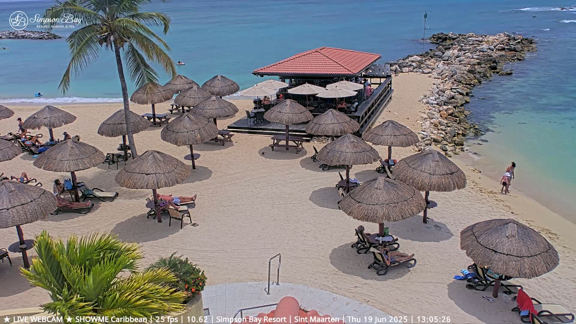 A sandy beach with numerous thatched umbrellas, beach chairs, and a beachfront bar, under a sunny sky and calm turquoise water.