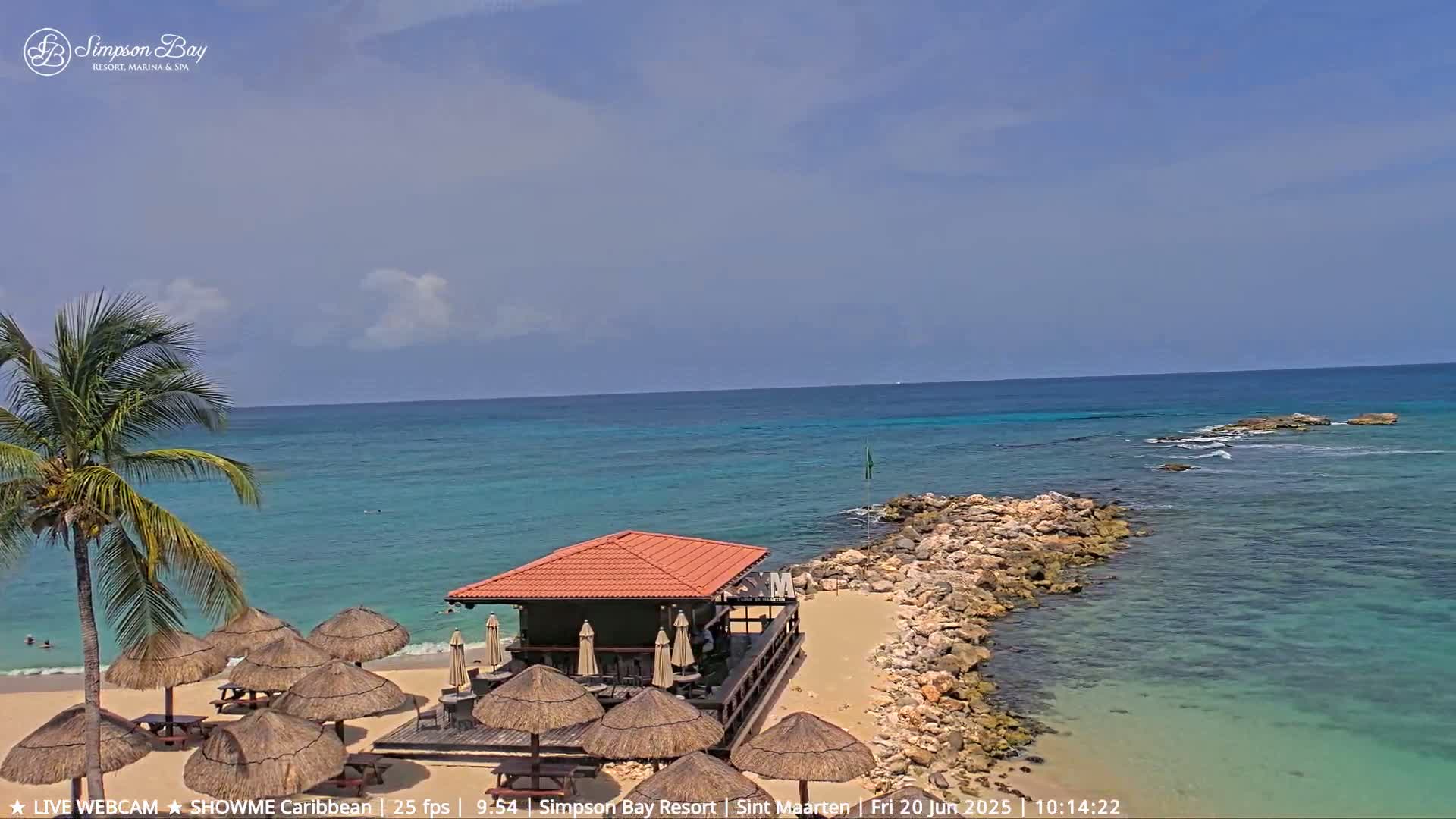 A beach scene with turquoise water, a small building with a red roof on a sandy shore, palm trees, and numerous thatched umbrellas under a partly cloudy sky.