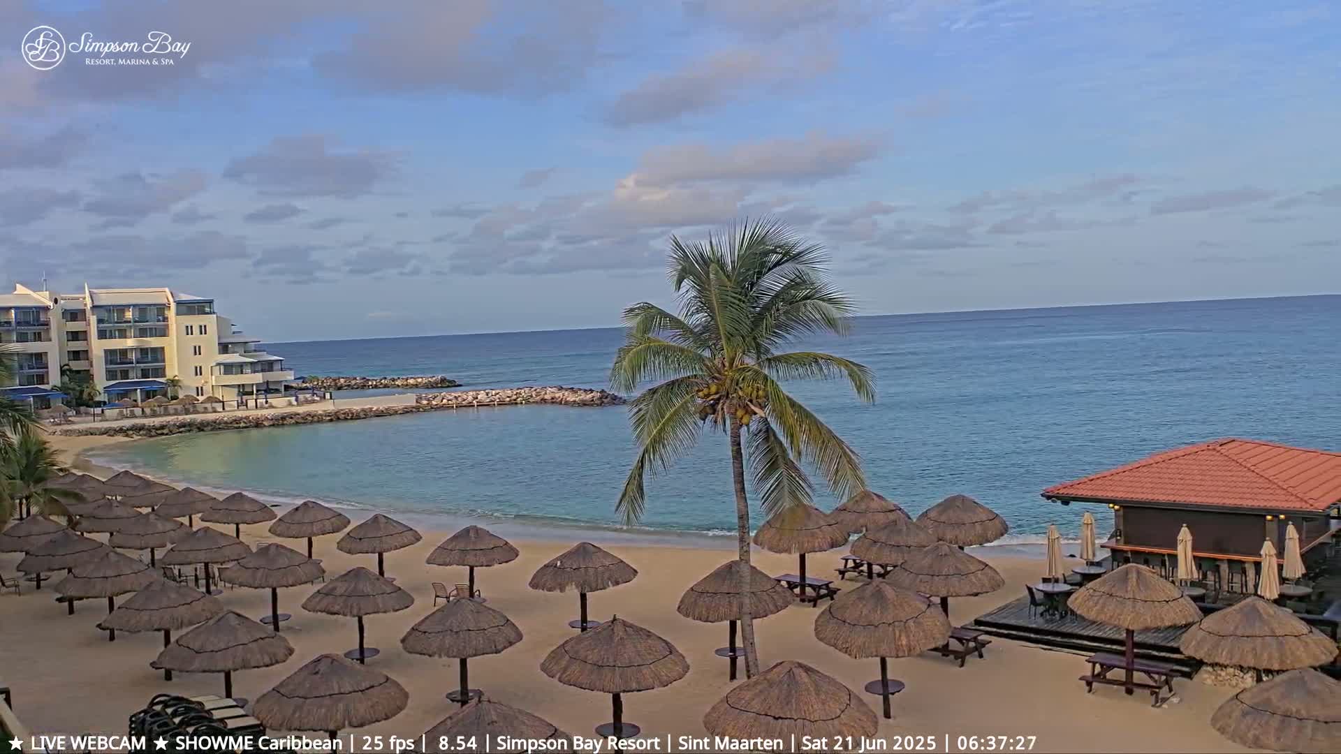 A sandy beach with numerous thatched umbrellas, a beachfront bar, a resort building, and a calm ocean under a partly cloudy sky.