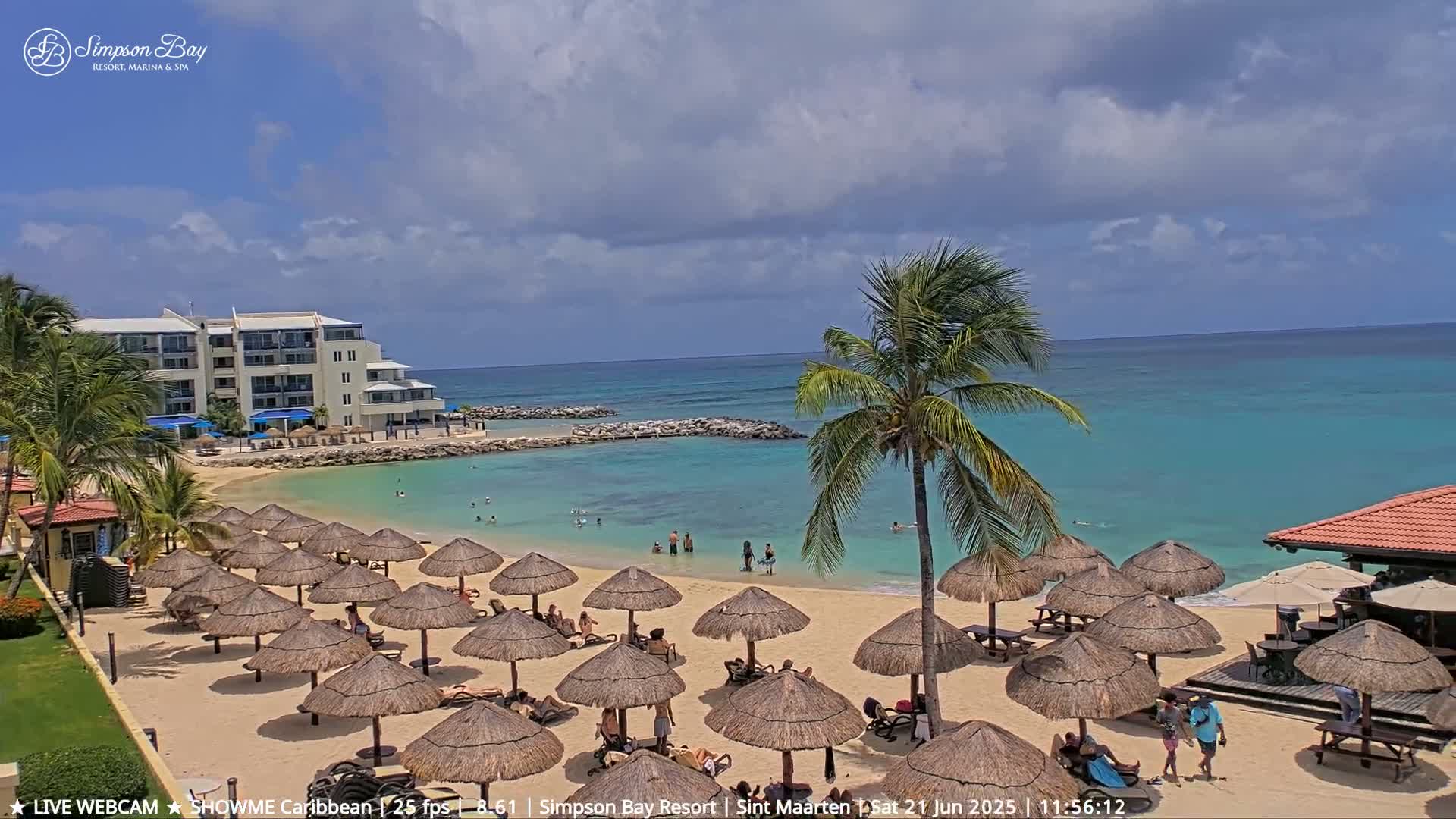 A sandy beach with numerous thatched umbrellas, beachgoers, and a resort building is situated next to a calm, turquoise ocean under a partly cloudy sky.
