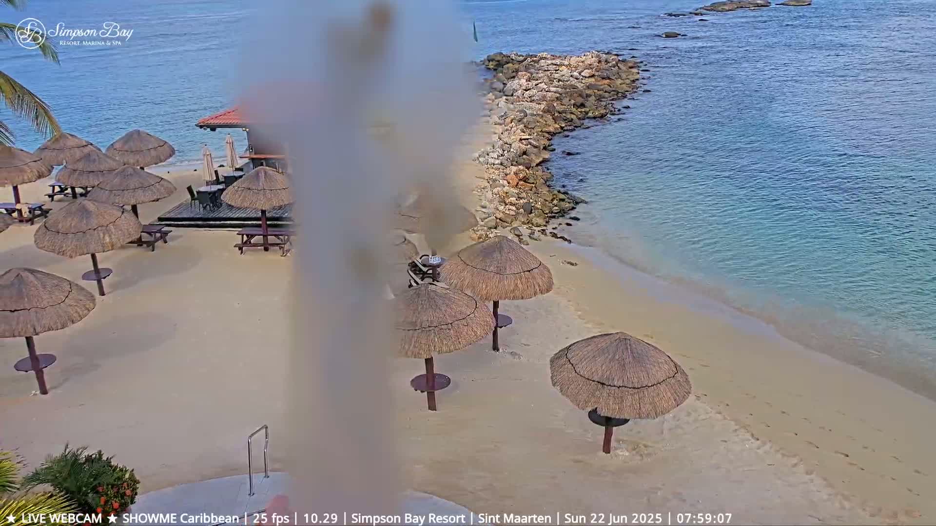 A sandy beach with thatched umbrellas and outdoor tables next to a calm, blue ocean, bordered by rocks.