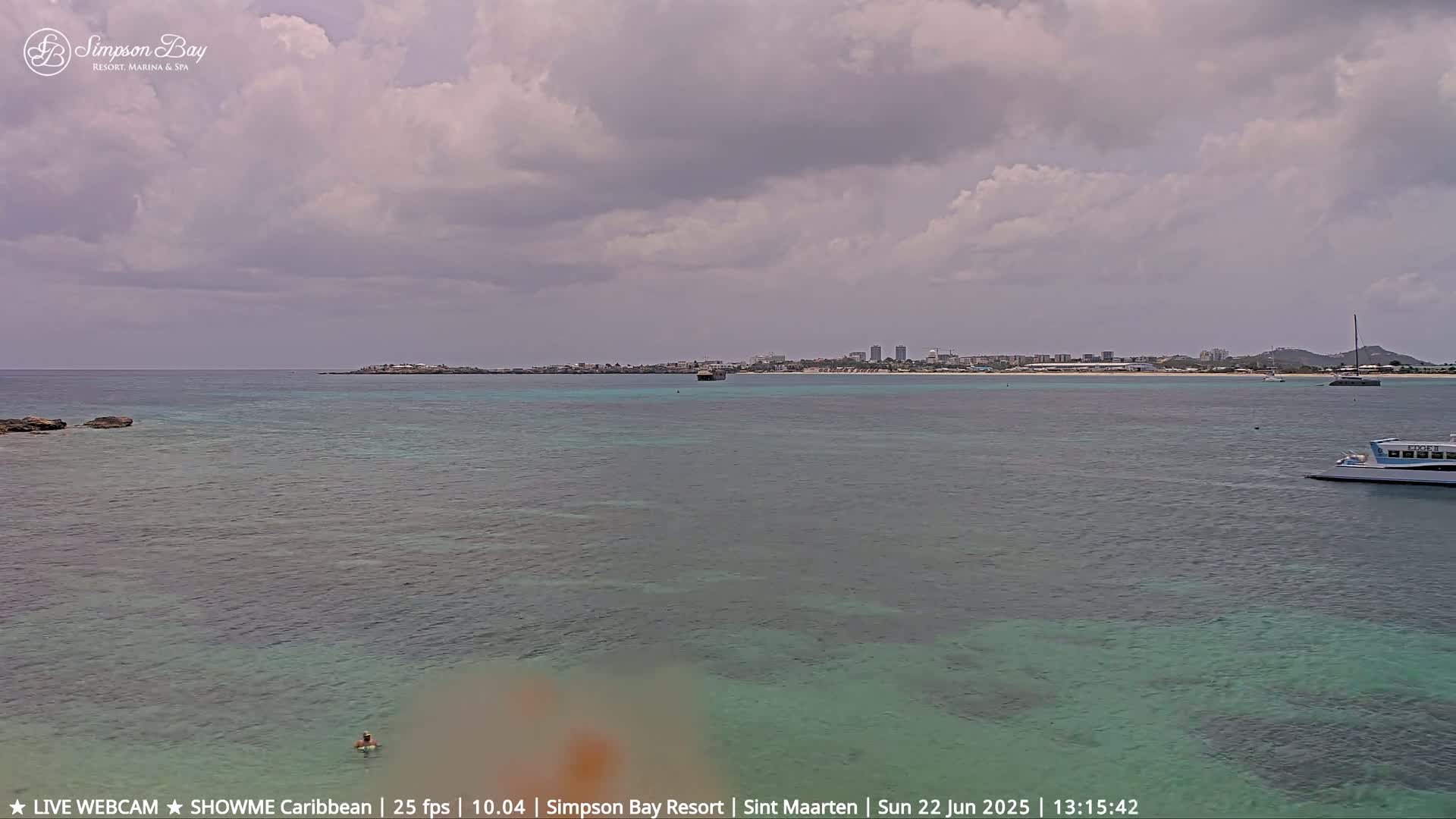 A partly cloudy day shows a calm, shallow turquoise ocean with a person swimming near the shore, boats in the distance, and a distant cityscape under a mostly cloudy sky.