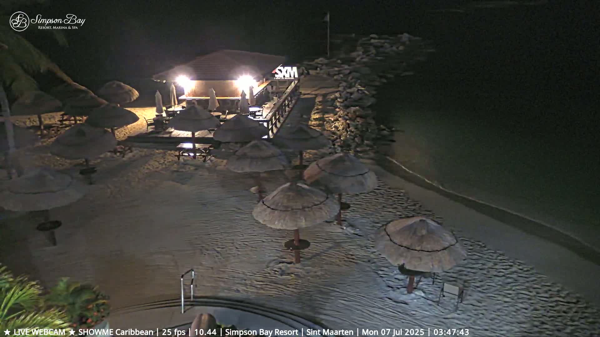 A nighttime beach scene shows a lighted bar and several thatched umbrellas on a sandy beach next to calm water.