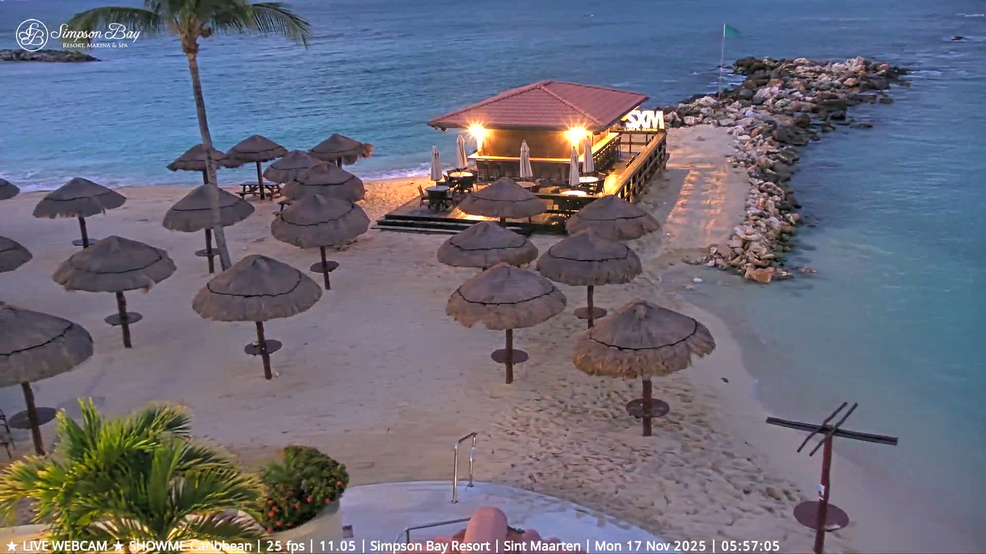 A serene tropical beach is shown at dusk or dawn with a brightly lit bar on a wooden deck, numerous straw umbrellas scattered across the sand, and a rock jetty extending into the calm, clear blue ocean under clear skies.