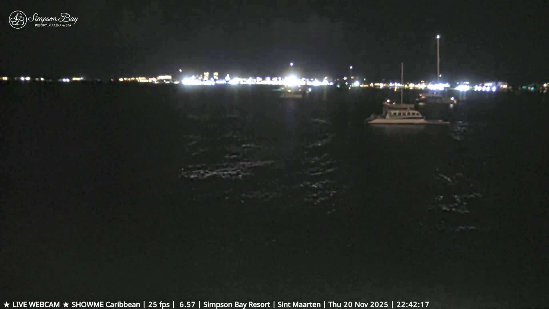A serene tropical beach is shown at dusk or dawn with a brightly lit bar on a wooden deck, numerous straw umbrellas scattered across the sand, and a rock jetty extending into the calm, clear blue ocean under clear skies.