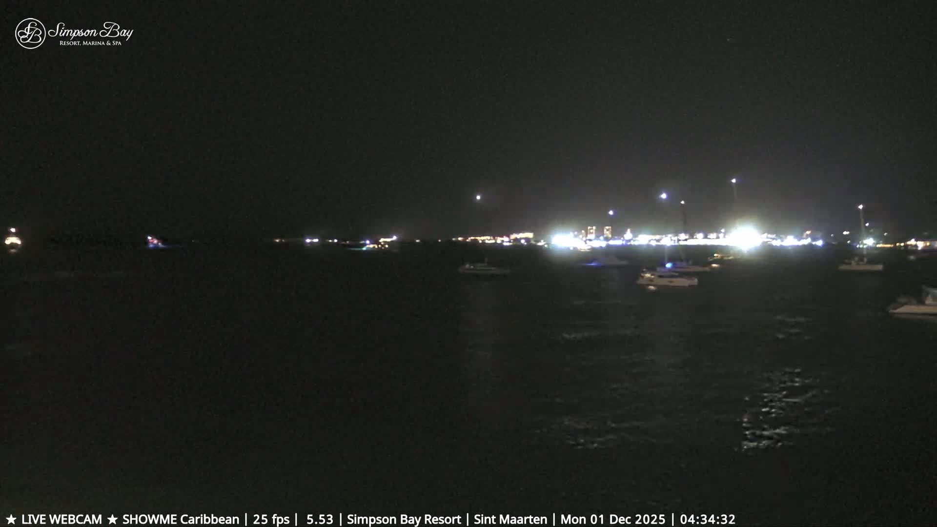 A clear night scene over a dark body of water reveals numerous boats with lights, a brightly lit distant shoreline with buildings, and the moon visible in the sky.
