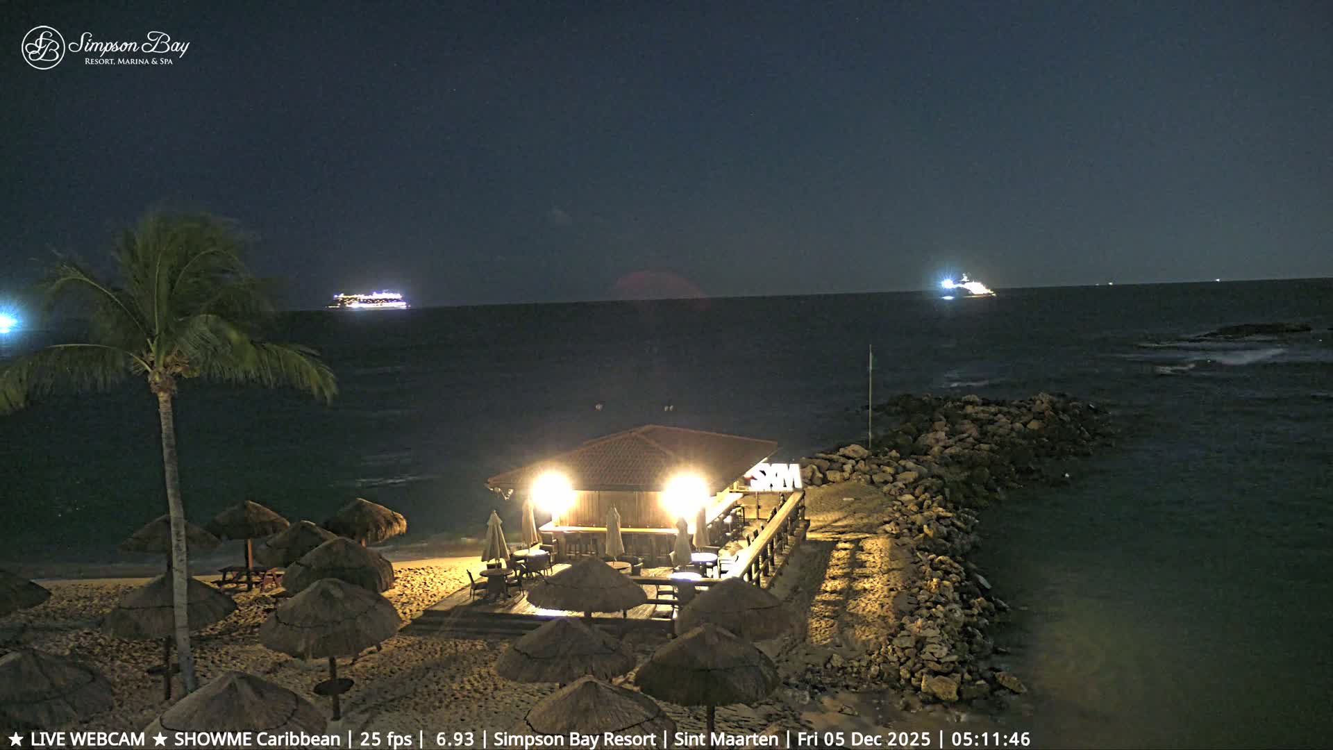 On a clear night, a brightly lit beach bar and straw umbrellas line the sand next to a palm tree and a rocky breakwater, with two illuminated ships visible on the dark ocean.