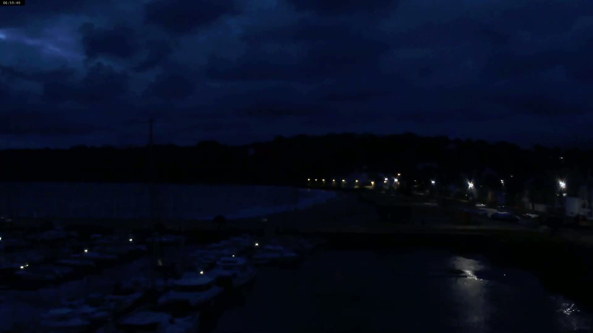 Under a dark, cloudy night sky, a body of water reflects the faint lights of a shoreline town with docked boats.
