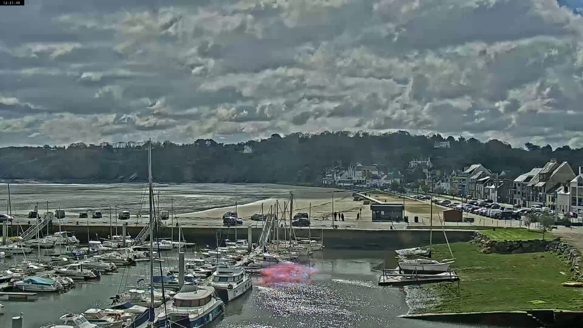 A marina filled with sailboats is situated next to a beach and a town under a partly cloudy sky.