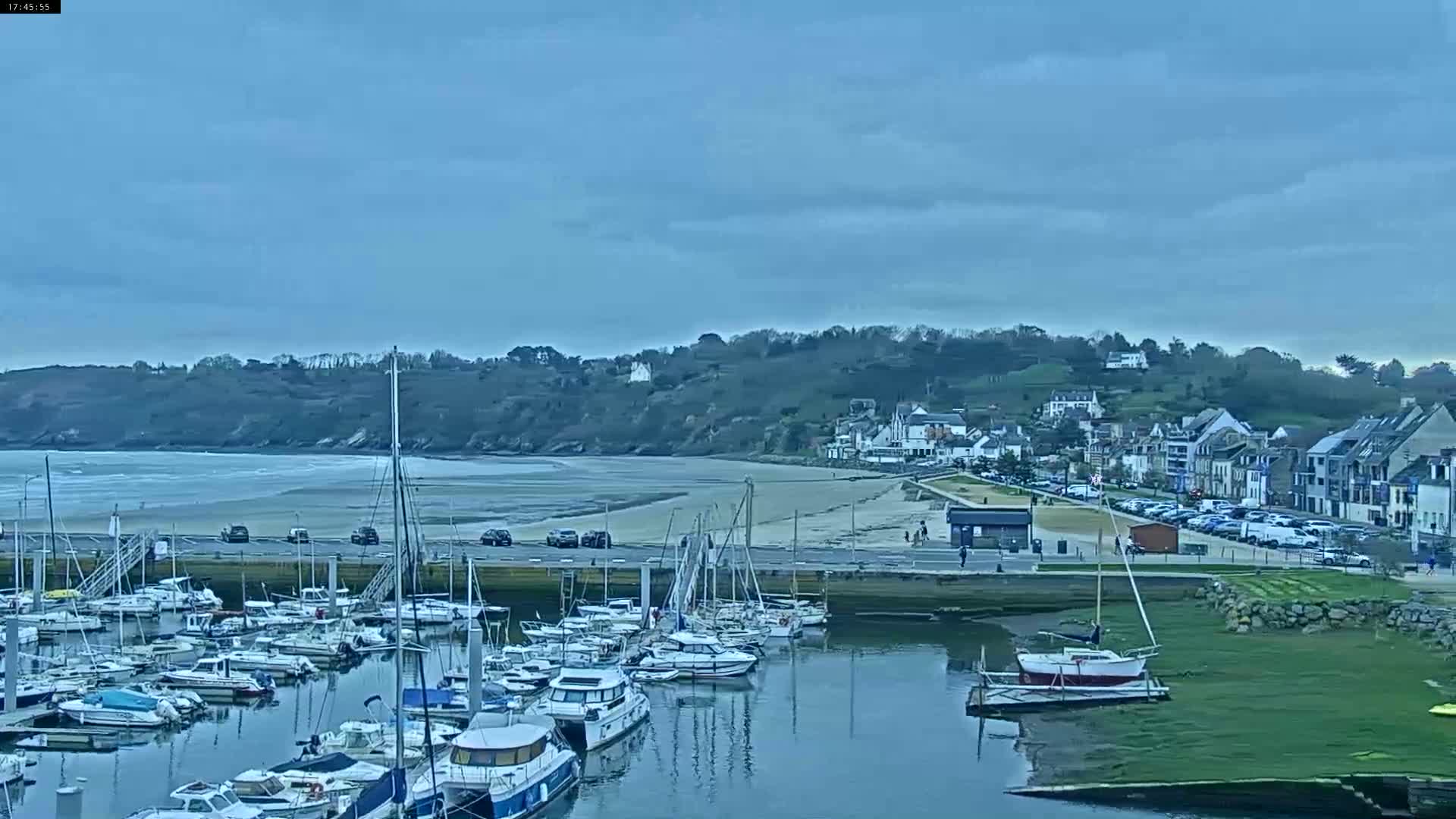 A marina filled with sailboats and motorboats is situated next to a sandy beach and a small town under a cloudy sky.