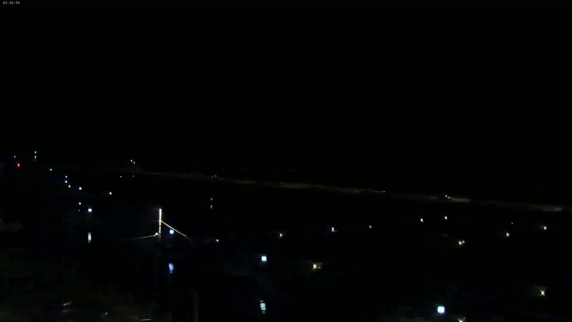 A dark nighttime scene shows a sparsely lit area, possibly a marina or dock, with faint lights scattered across the water and along a shoreline.