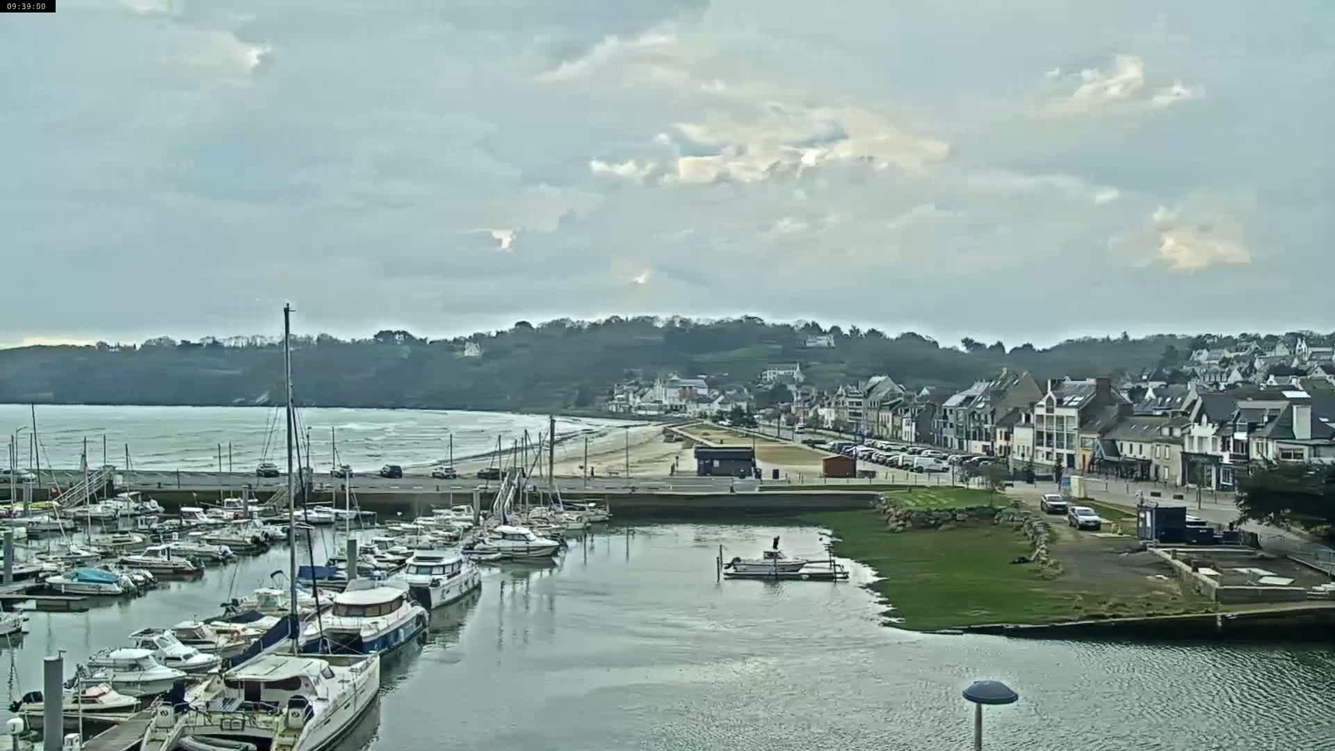 A harbor filled with sailboats and motorboats is situated next to a town with a sandy beach under a mostly cloudy sky.