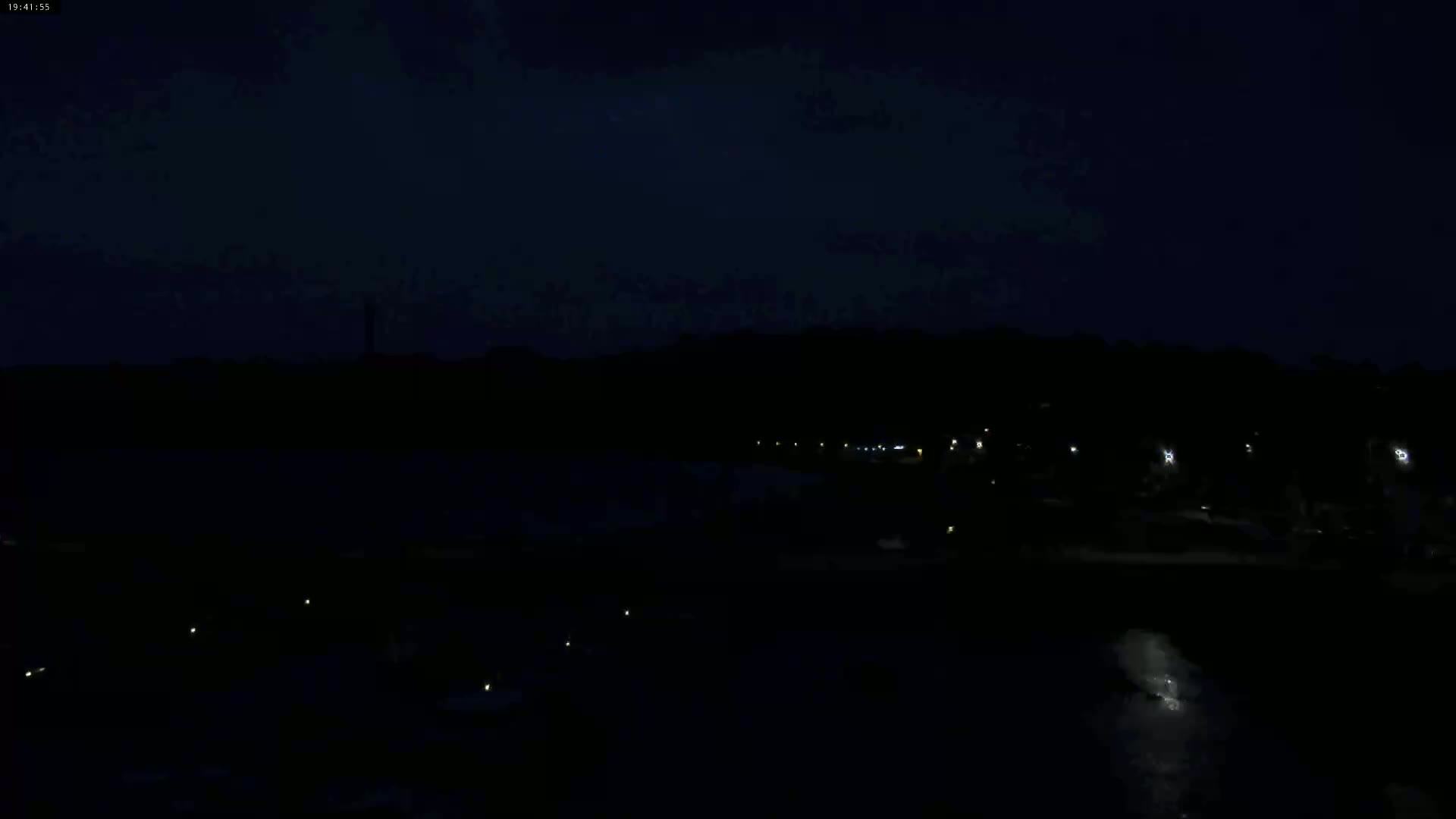 A dark nighttime scene shows a body of water with a few faint lights along the shore, and a darker landmass with more lights visible in the distance under a mostly clear, dark sky.
