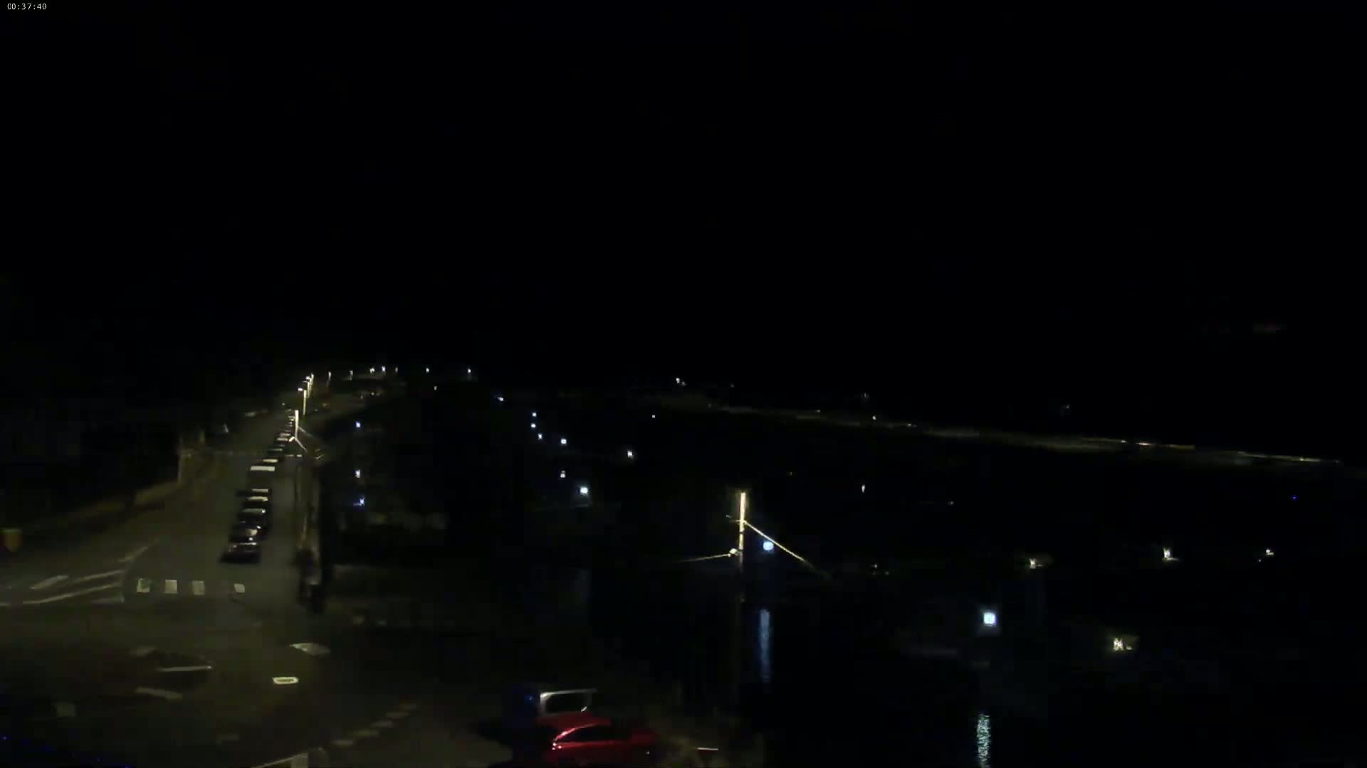 A nighttime view shows a road with cars and streetlights leading to a dark body of water with faint lights along its edge.