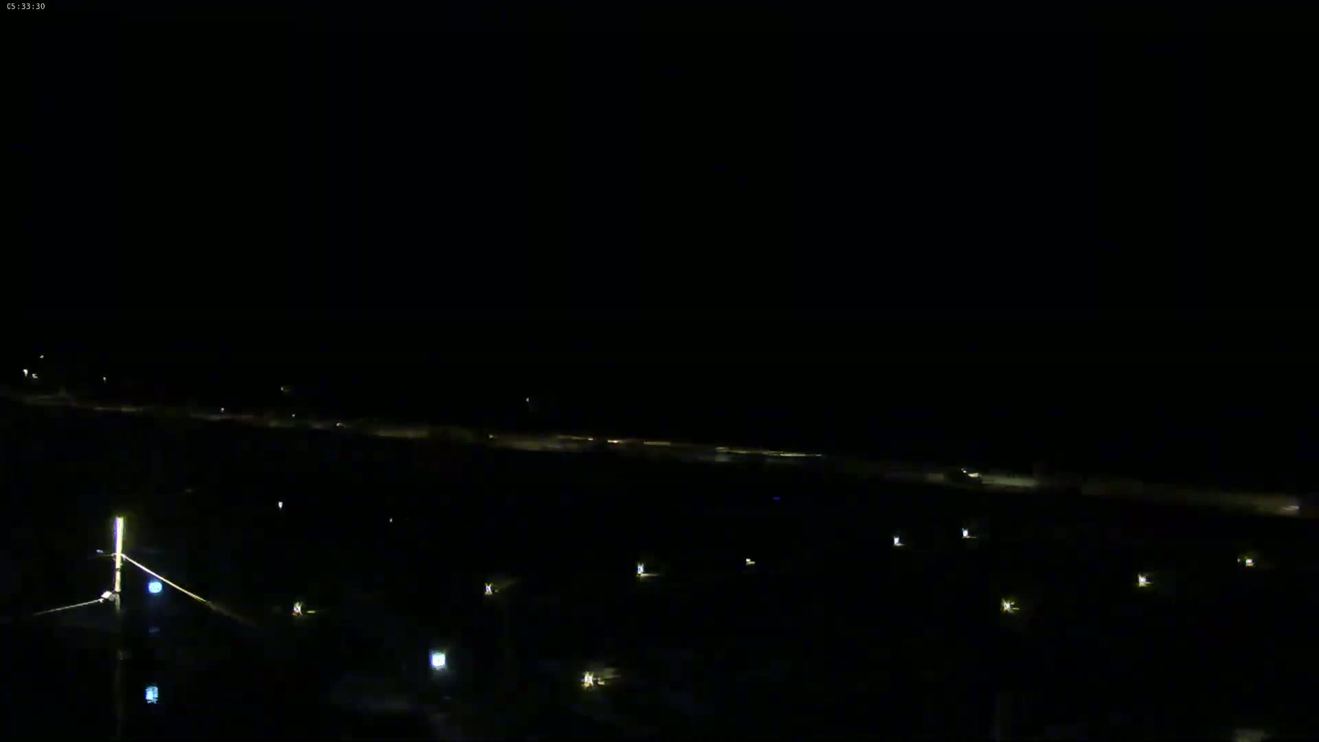 A nighttime, long-range view shows a dark, flat expanse of land with a line of distant lights, possibly a road or shoreline.