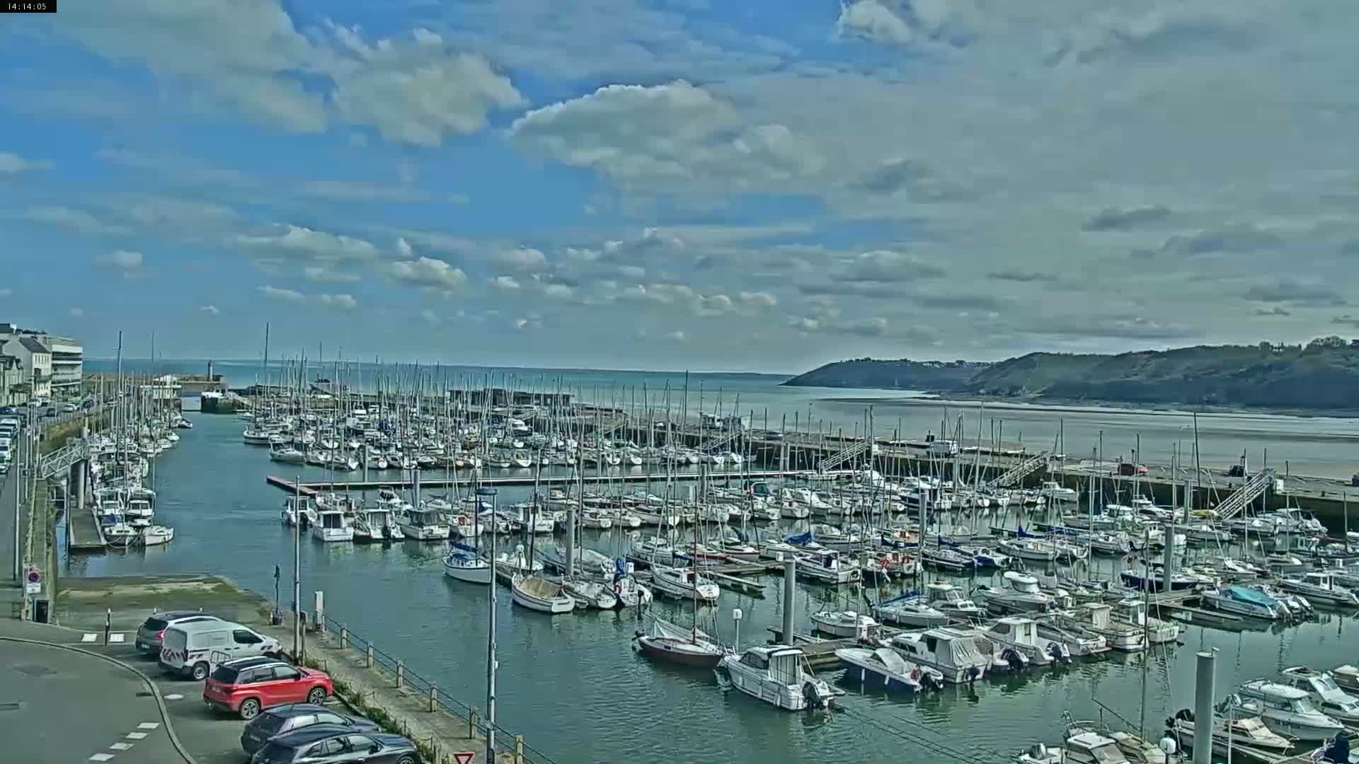 A marina filled with numerous sailboats and motorboats is situated on a calm body of water under a partly cloudy sky.