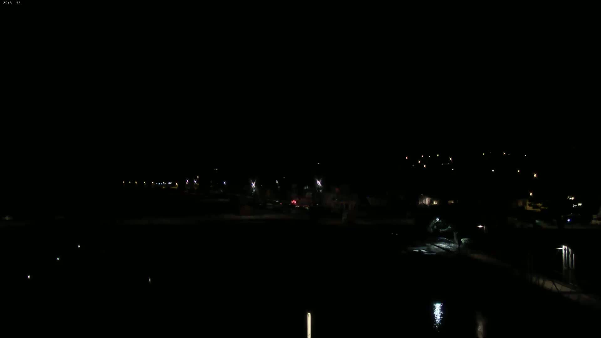 A dark nighttime scene shows a sparsely lit waterfront area with a few distant lights on a hill in the background.