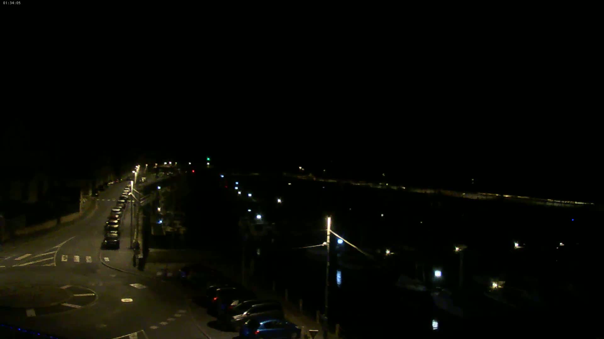 A nighttime, high-angle view shows a road with parked cars and streetlights, leading to a dark body of water with faintly illuminated boats.