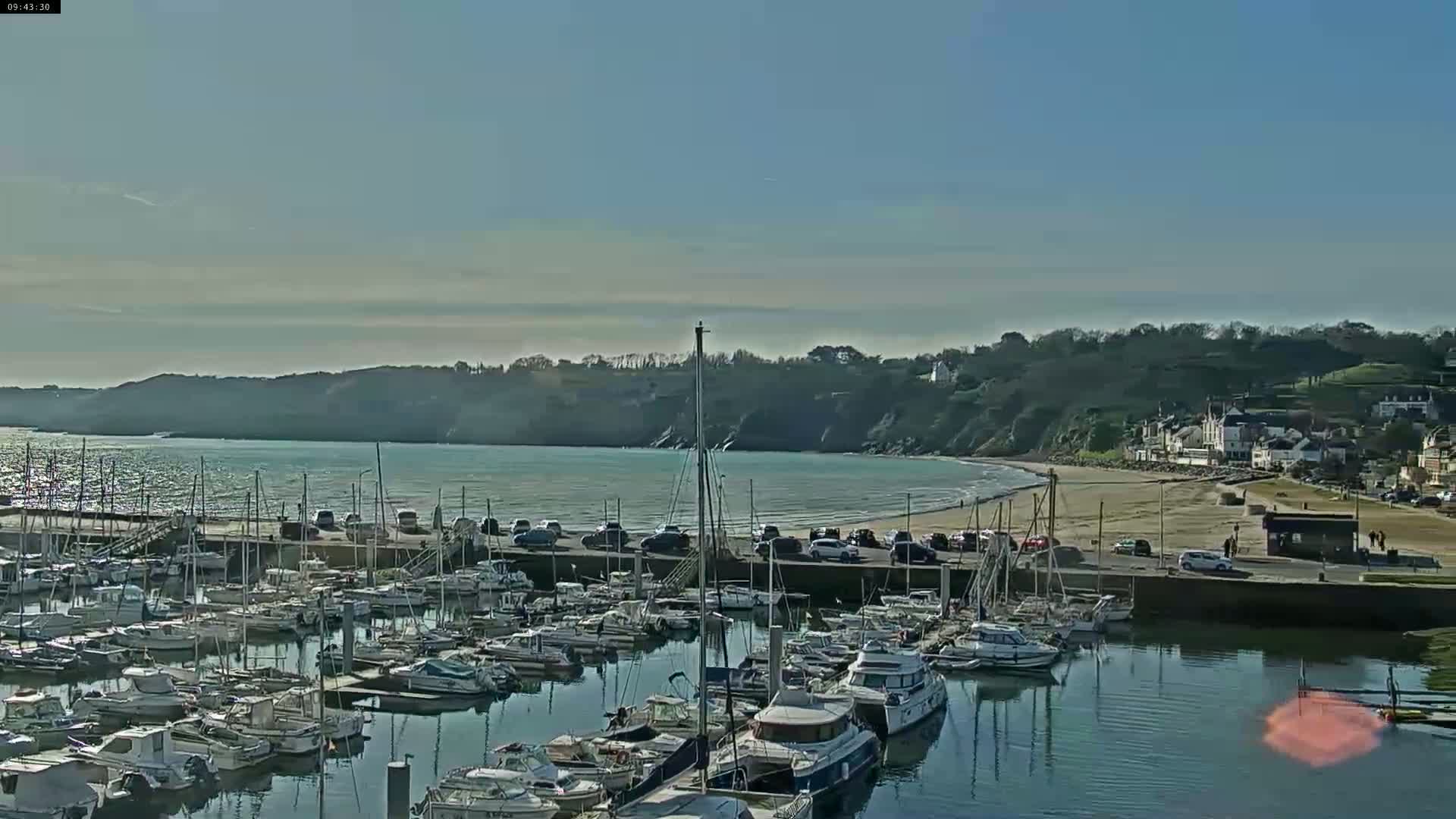 A marina filled with sailboats and motorboats is situated next to a calm bay with a sandy beach and a small town nestled on a hillside under a clear, sunny sky.