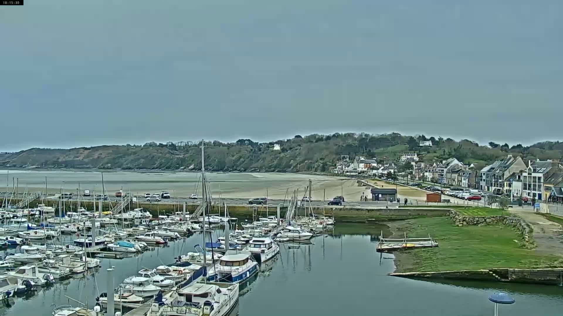 A harbor filled with numerous sailboats and motorboats is situated next to a sandy beach and a small town under a mostly cloudy sky.