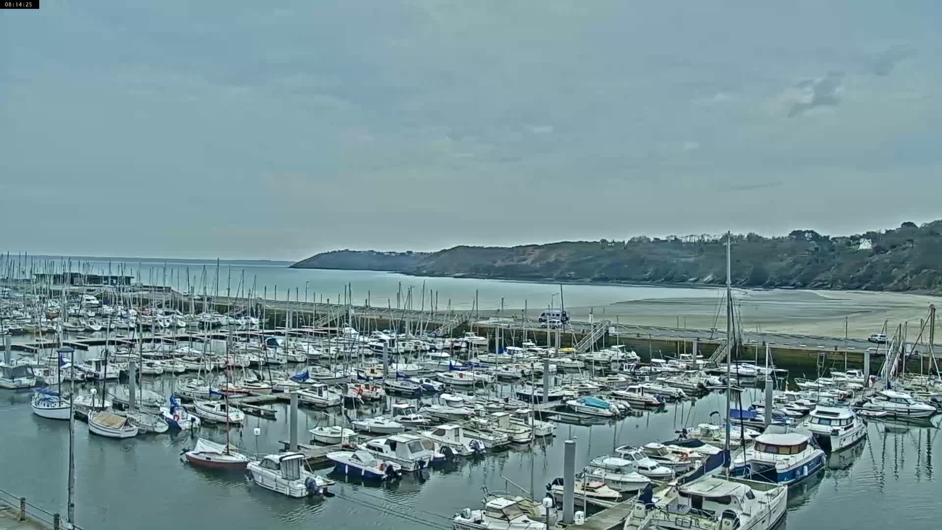 A marina filled with numerous sailboats and motorboats is situated along a calm bay under a cloudy sky.