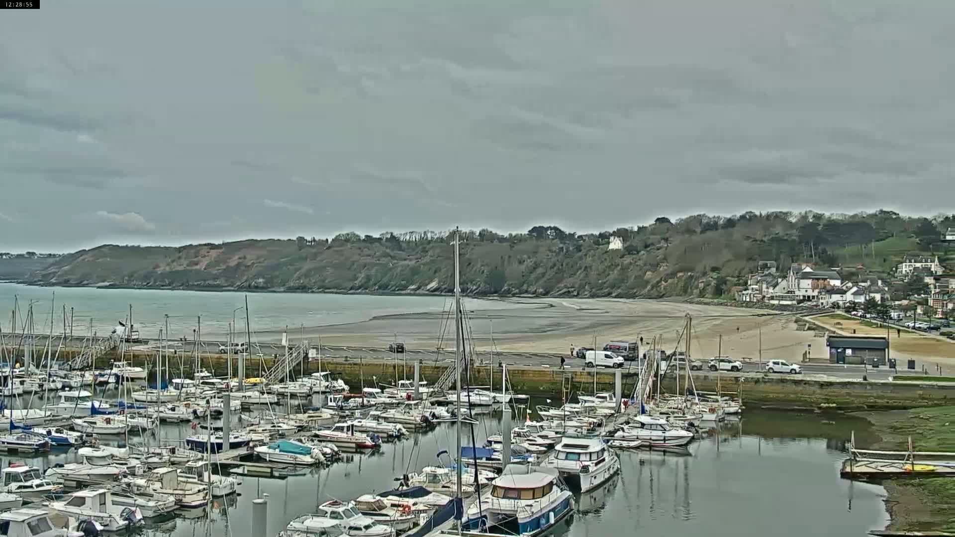 A marina filled with numerous boats is situated next to a sandy beach and a low, tree-covered hillside under a cloudy sky.