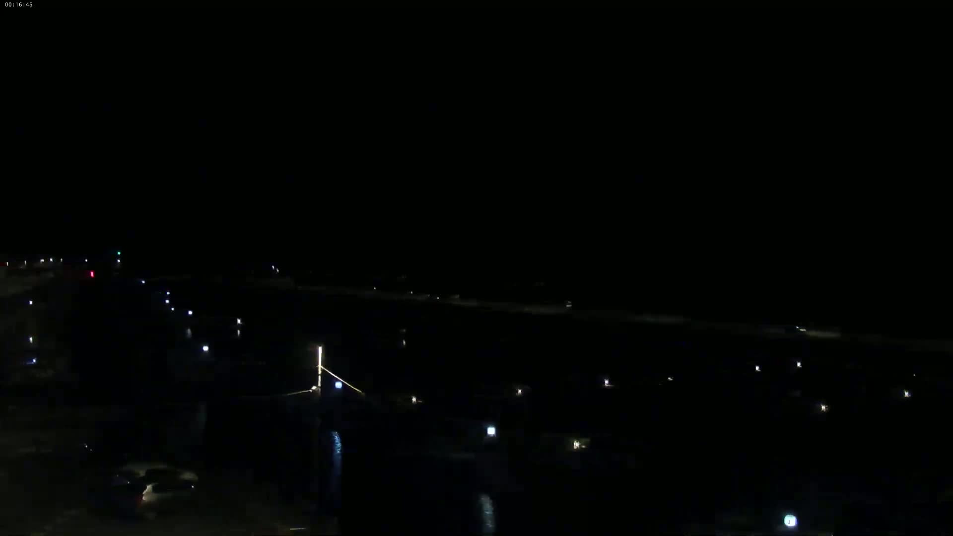 A nighttime, dark scene shows a long, sparsely lit roadway and a body of water with faint lights visible in the distance.