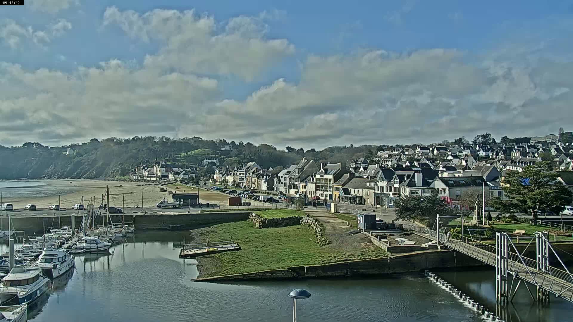 A coastal town with a marina filled with boats is seen under a partly cloudy sky, with a sandy beach visible in the distance.