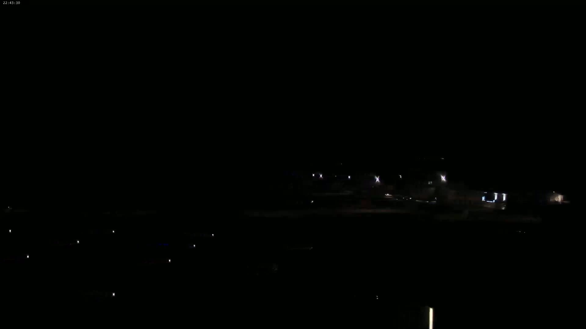 A dark, nighttime scene shows a few distant lights from buildings and vehicles, under a clear, black sky.