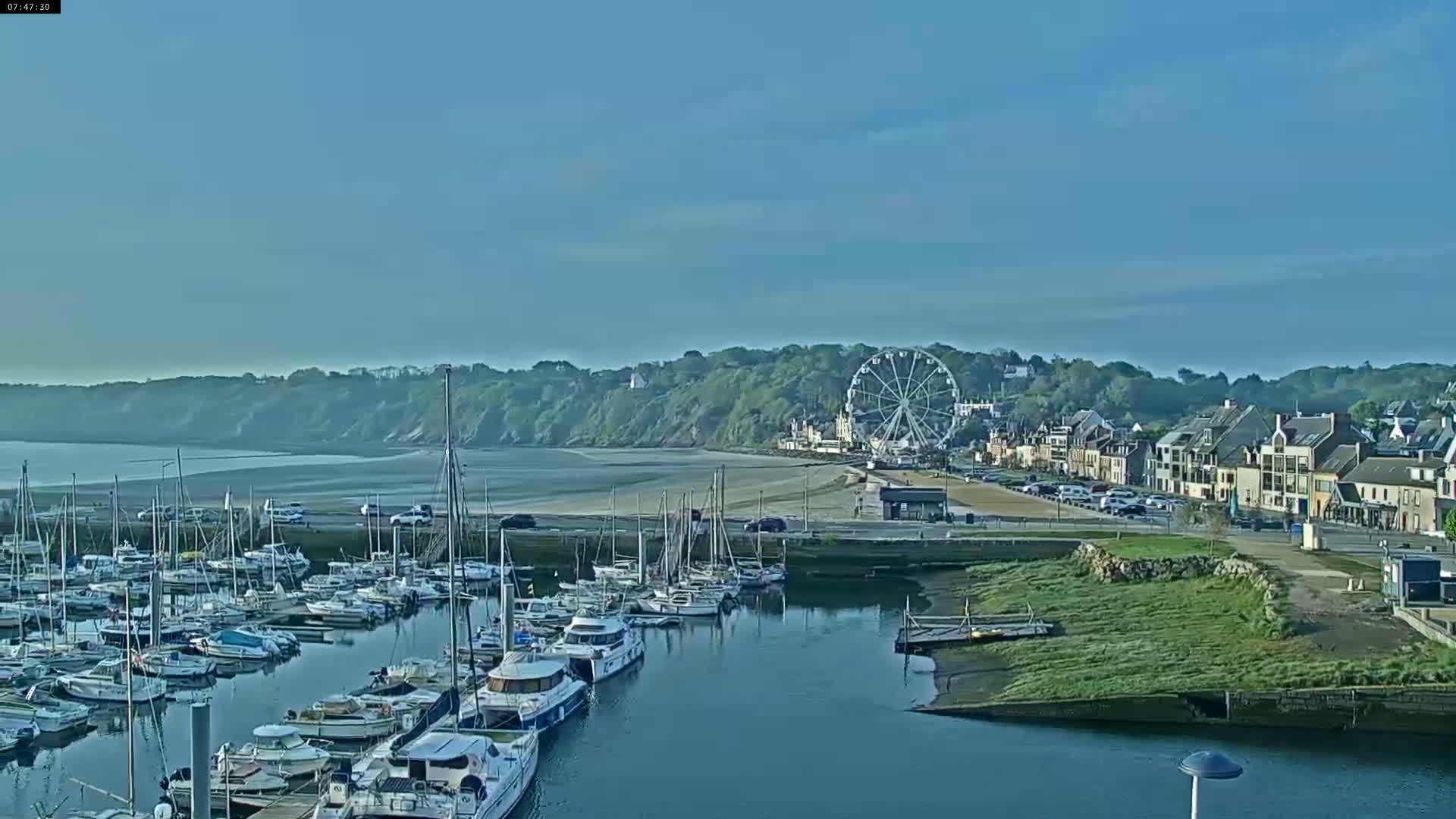 A marina filled with sailboats and motorboats is situated near a town with a Ferris wheel on a clear, sunny day.