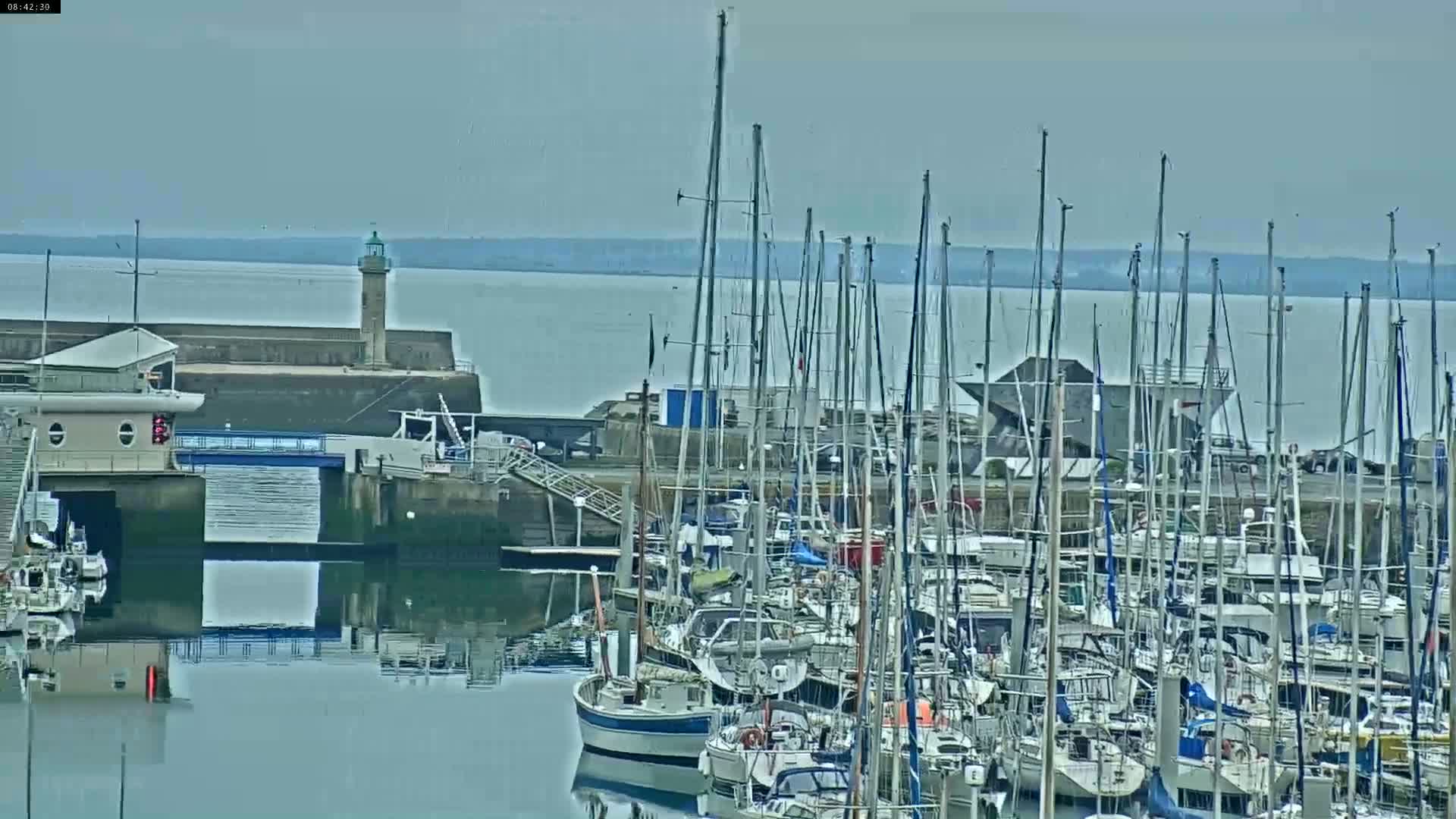 Many sailboats are docked in a harbor on a calm, overcast day, with a lighthouse visible in the distance.