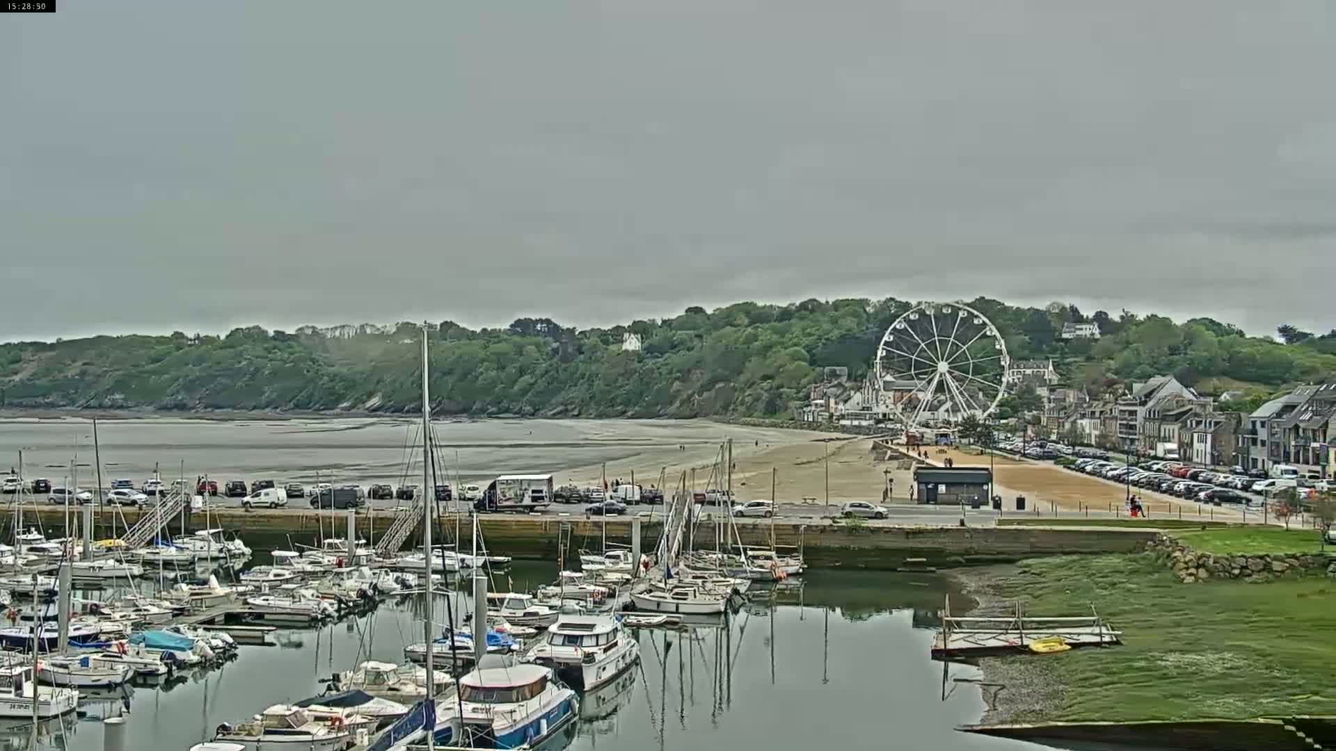 A marina filled with sailboats and motorboats is situated next to a sandy beach, beyond which sits a Ferris wheel and a line of buildings on a hillside under an overcast sky.