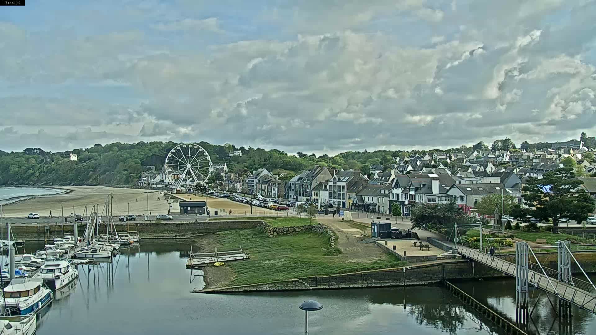 A seaside town with a Ferris wheel, harbor filled with boats, and a sandy beach under a partly cloudy sky.