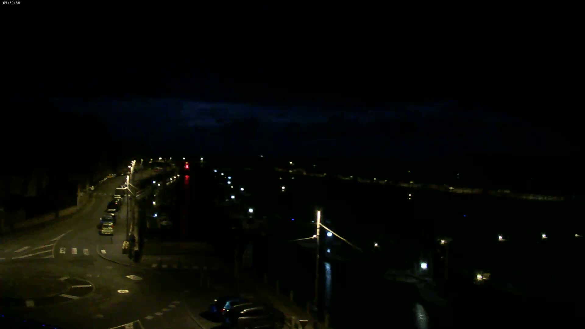 A nighttime, dimly lit view of a road intersecting near a waterway with sparsely lit boats and buildings along the shore under a dark, cloudy sky.