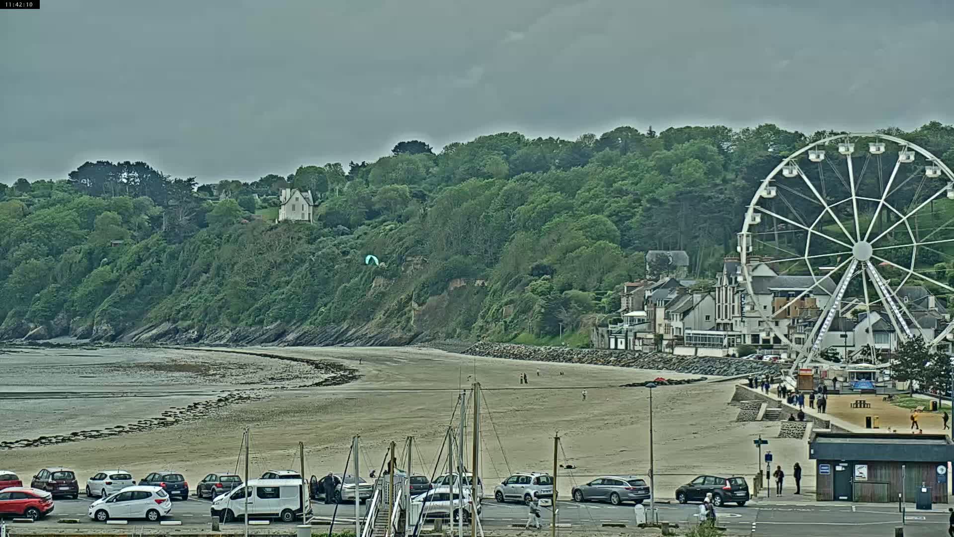 A sandy beach with a Ferris wheel and parked cars, backed by a green hill and overcast sky.