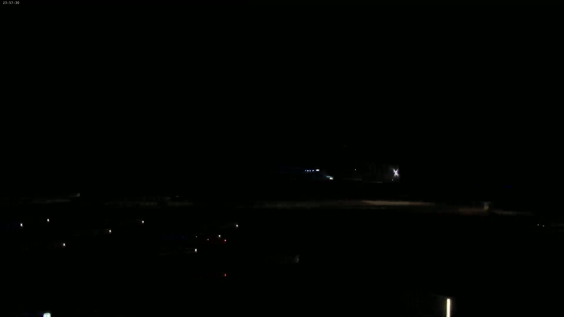 A nighttime, dark scene shows a few distant lights in the mid-ground and several more faint lights below, likely vehicles, on a mostly empty expanse.