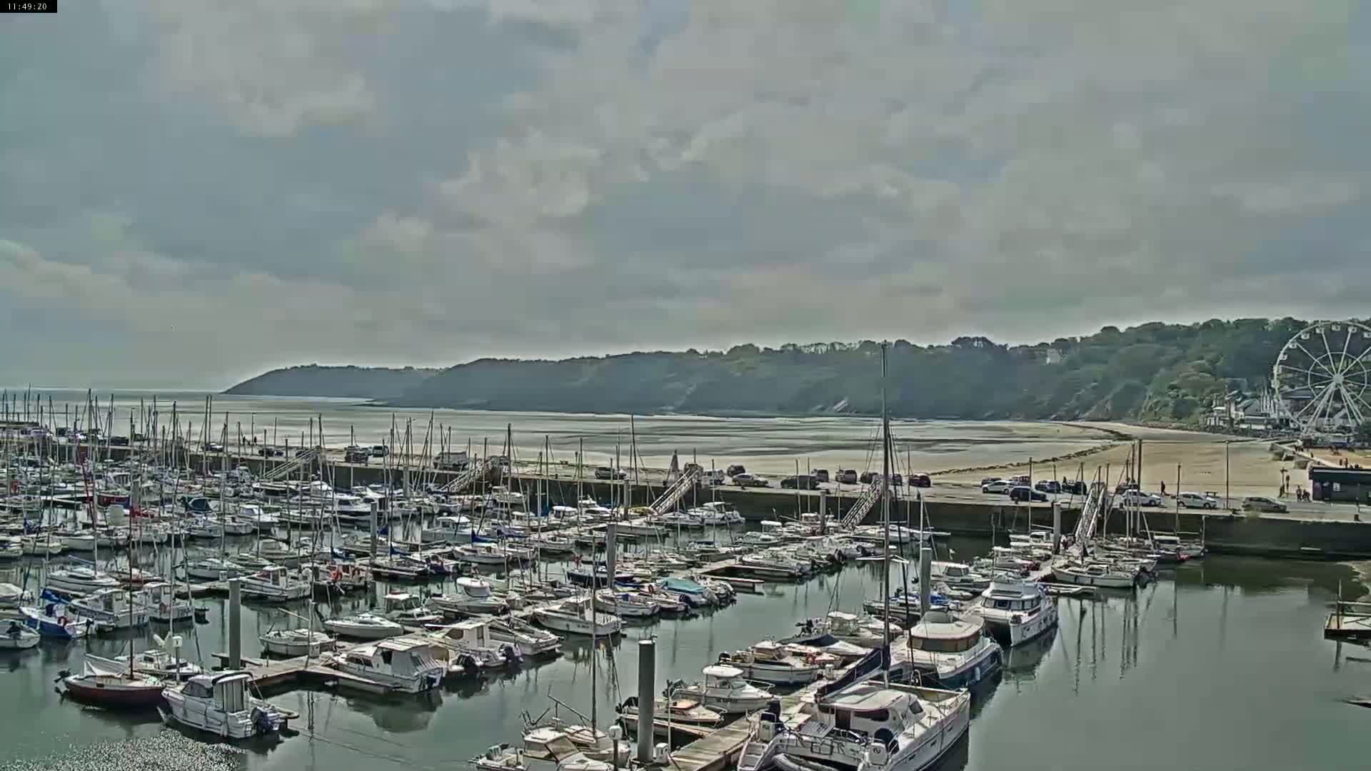 A harbor filled with numerous sailboats and motorboats is situated near a sandy beach and a Ferris wheel under a partly cloudy sky.