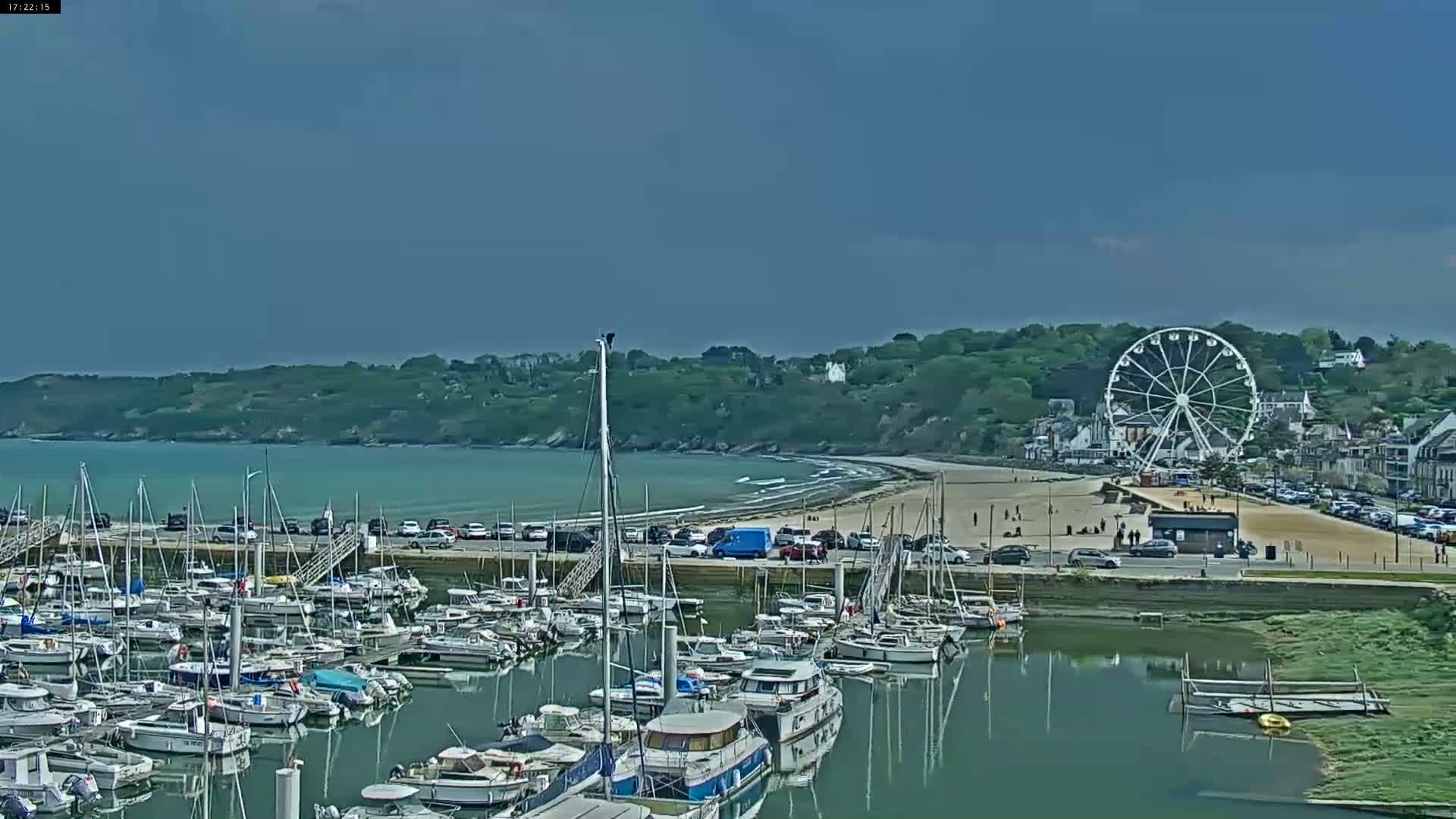 A marina filled with numerous boats is situated near a beach with a Ferris wheel, all under a partly cloudy sky.