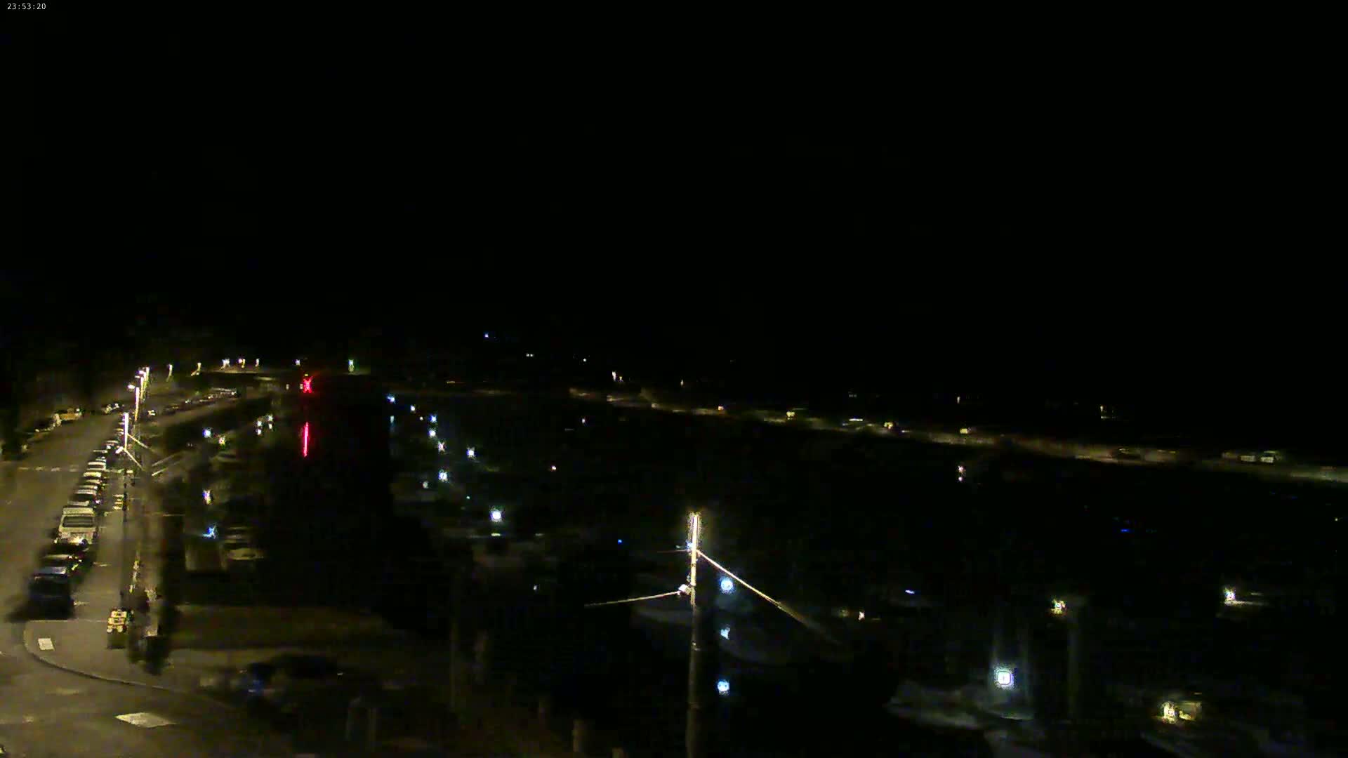 A nighttime view of a harbor with parked cars along a roadway and lights illuminating the scene.