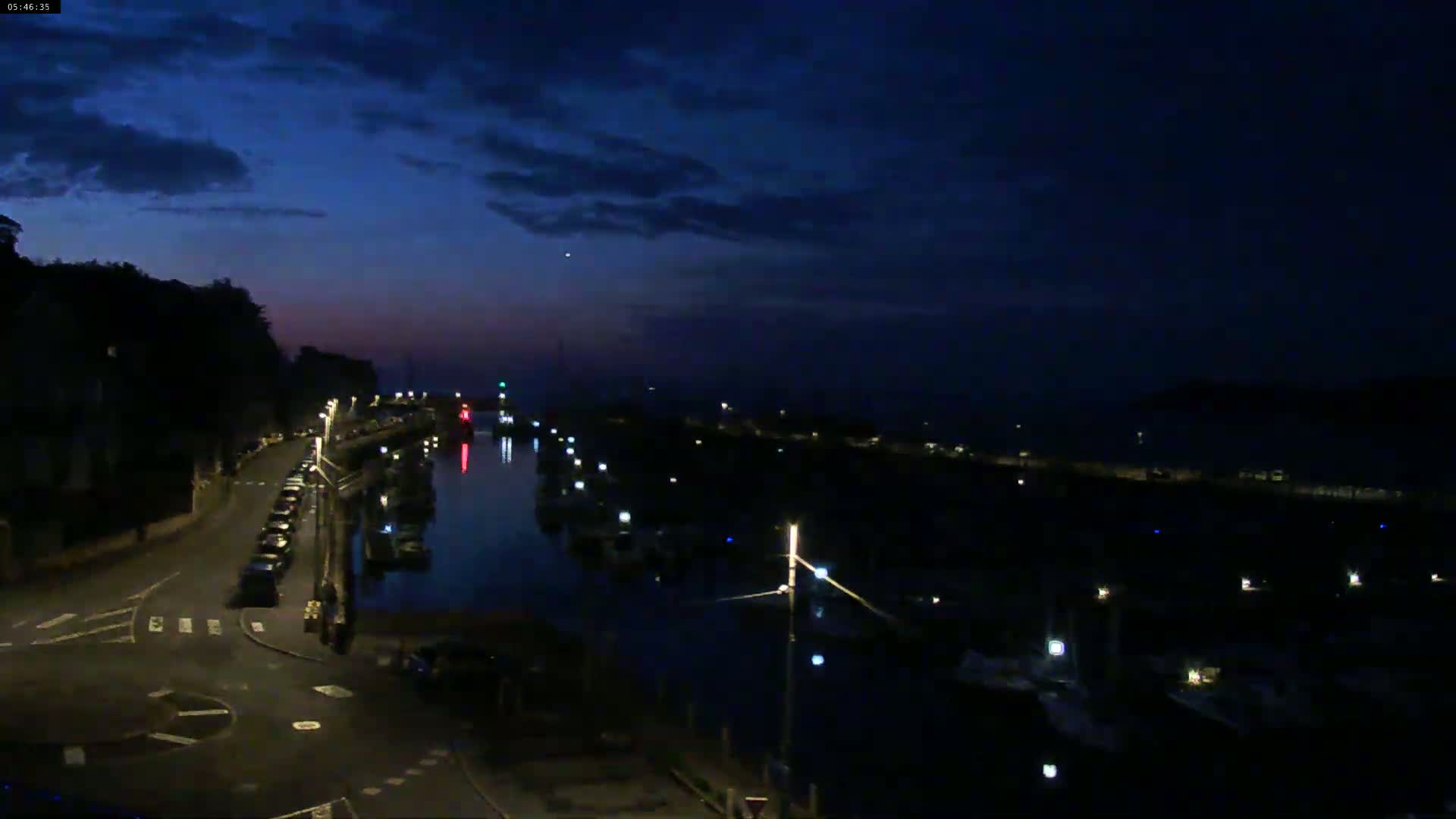 A dark, nighttime view of a harbor filled with boats, a road with parked cars, and a hill in the background under a mostly cloudy sky.
