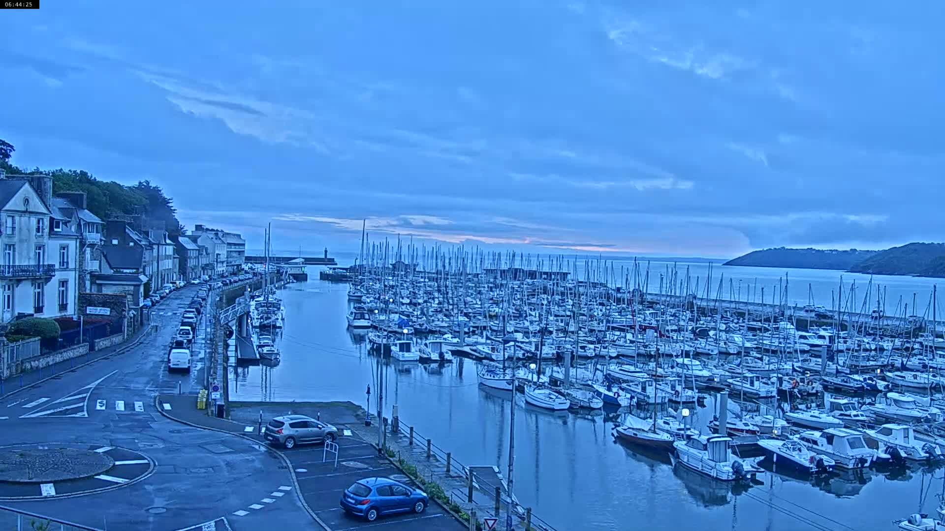 A harbor filled with numerous sailboats and motorboats is seen under a partly cloudy, blue sky at dawn, with a town and road visible in the foreground.