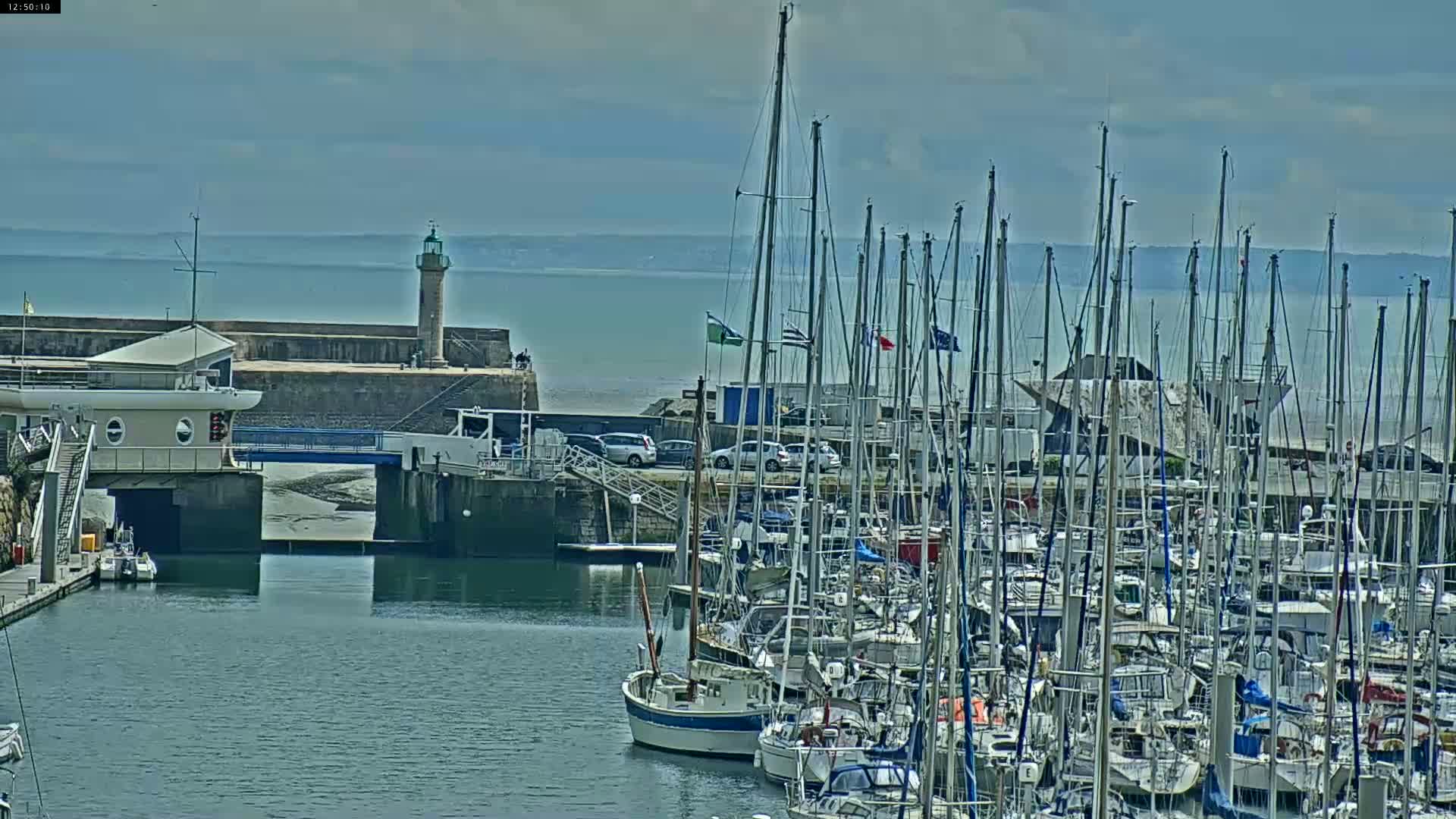 A marina filled with numerous sailboats is situated next to a stone harbor wall with a lighthouse under partly cloudy skies.