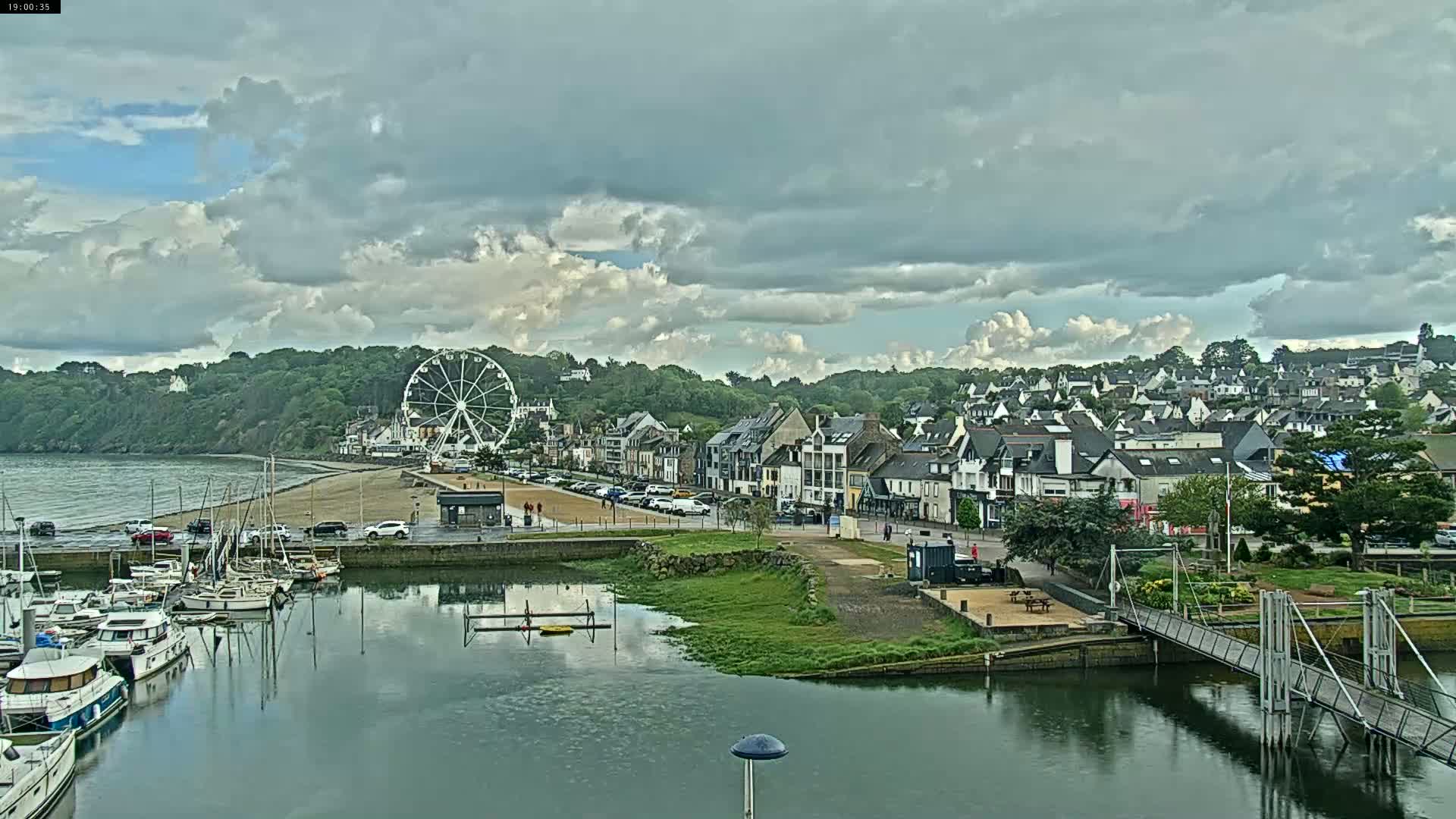 A harbor filled with boats sits below a coastal town with a Ferris wheel visible on the beach under a cloudy sky.