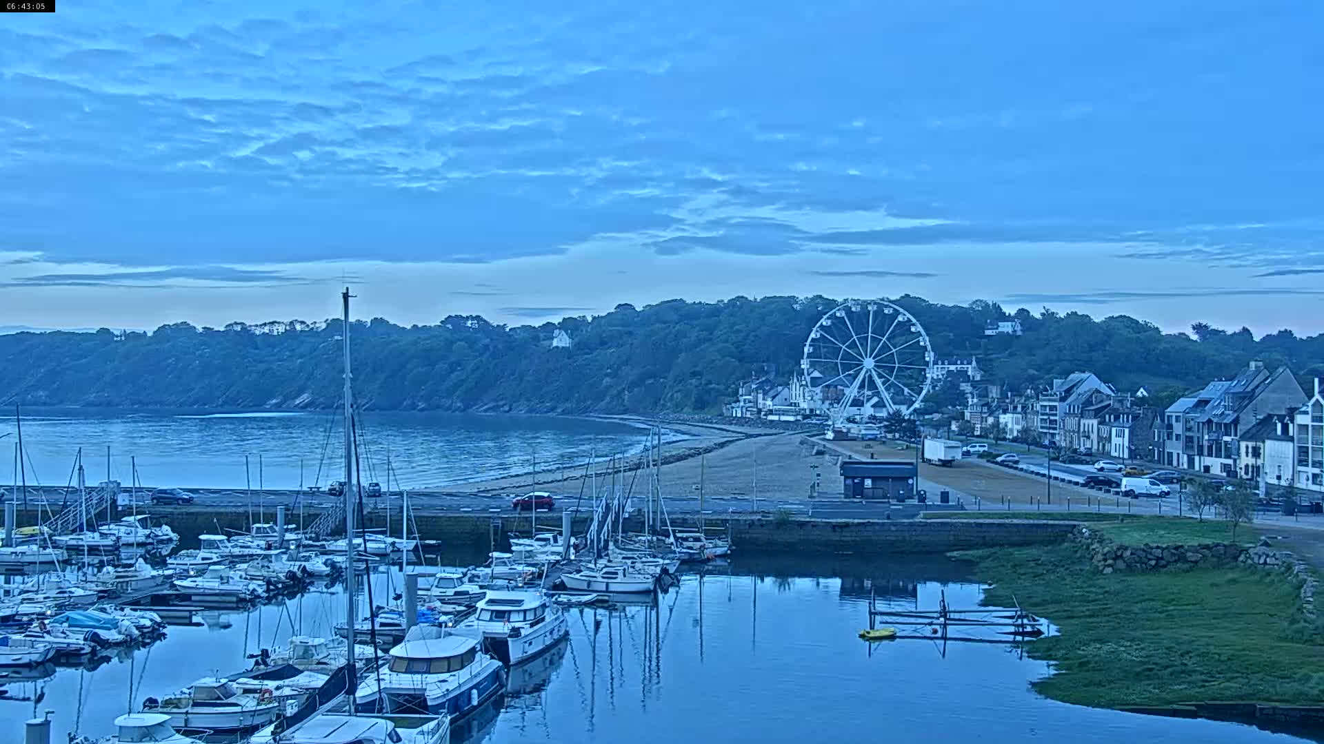A marina filled with sailboats and motorboats is situated below a hill with buildings and a Ferris wheel, all under a twilight sky.