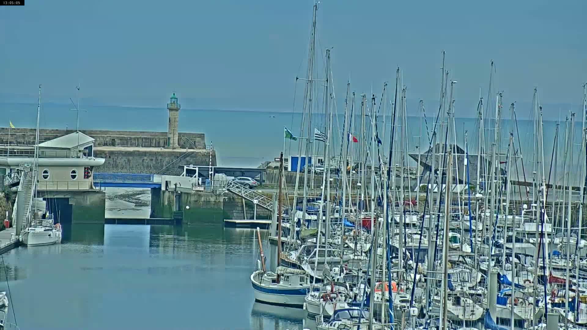 A marina filled with numerous sailboats is situated next to a stone harbor wall with a lighthouse under a clear, sunny sky.